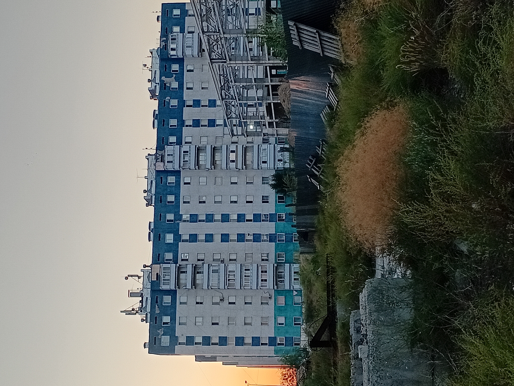 The image depicts a large, multi-story apartment building in the city of Semey, Kazakhstan. The building has a light-colored facade with horizontal bands of blue on the upper floors and a turquoise band on the lower floors. Numerous windows and balconies are visible. The roof of the building is equipped with antennas and what appear to be ventilation shafts.
To the right of the apartment building, a steel frame structure, likely under construction, is prominent. It is a skeletal framework of beams and columns, suggesting a new building or expansion project.
In the foreground, the scene transitions to an undeveloped area with overgrown vegetation, including tall grasses and bushes. There are also piles of construction debris, including concrete blocks, wooden pallets, and corrugated metal fencing. The lighting suggests it is either early morning or late evening, with a soft, warm glow on the left side of the image, indicating the position of the sun. No people are clearly visible in the scene. The overall impression is one of an urban environment undergoing development, with a mix of completed residential structures and ongoing construction amidst a somewhat unkempt foreground.