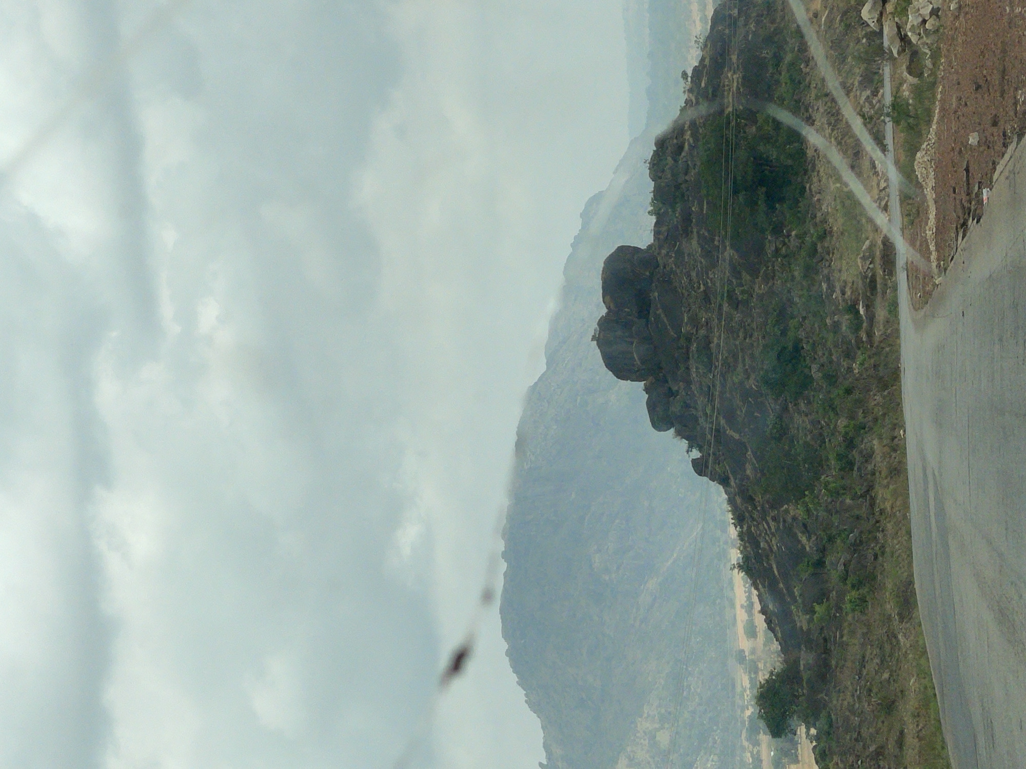 The image depicts a mountain landscape with a road running along the right edge of the frame. The road surface appears to be paved. Adjacent to the road is a steep, rocky hillside densely covered with vegetation. In the background, layers of mountains recede into the distance, obscured by a hazy atmosphere. The sky is overcast with clouds, creating diffused lighting conditions. There is no visible evidence to confirm the precise location, but the presence of rocky terrain suggests a location in the city of Vom, Nigeria. There are no visible people in the image.