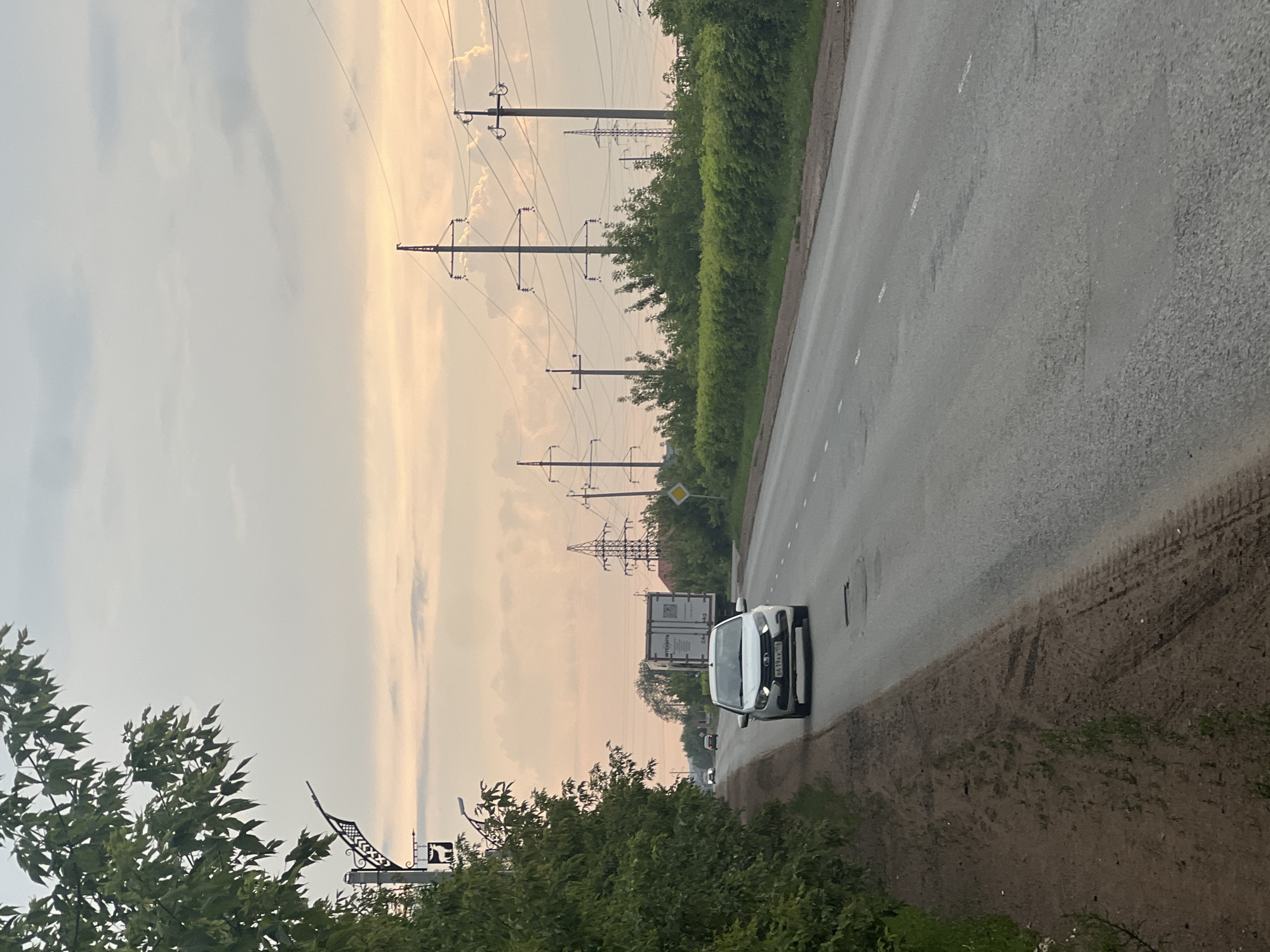This is a vertical, outdoor photograph taken from a slightly elevated perspective looking down a paved road. The sky dominates the upper portion of the image, with soft, diffused light suggesting either dawn or dusk. Large cumulus clouds are visible, interspersed with areas of lighter sky, hinting at overcast or humid weather. A line of tall power transmission towers runs along the left side of the road, with numerous wires stretching between them. Lush green trees and bushes line the edge of the road on the left, creating a natural border.
On the road, a white car is moving away from the camera. Further down the road, another white vehicle, possibly a truck or a larger car, is also visible, along with a smaller, distant vehicle. The road surface appears to be asphalt, with visible tire tracks and some patches of wear. The right side of the road is bordered by a dirt shoulder, also showing tire marks and some sparse vegetation.
In the foreground on the left, part of a tree with green leaves is visible, partially obscuring a decorative metal structure with a pointed roof and a sign.
The scene appears to be a rural or semi-rural setting in Buzuluk, Russia, likely during the daytime due to the overall brightness, though the light quality suggests either early morning or late evening. There are no discernible people in the image. The overall mood is calm and serene, capturing a moment of travel on a quiet road. A license plate on the white car is partially visible and reads "6619AX".