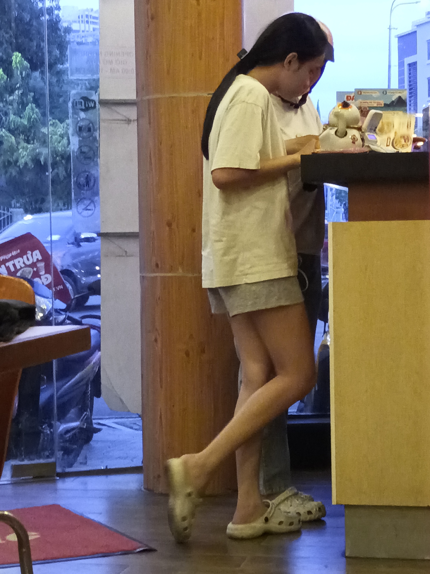 In Nha Trang Ward, Vietnam, a woman in a white t-shirt and grey shorts stands facing a counter. She is wearing white crocs and her dark hair is pulled back. She is looking down at the counter and appears to be holding something. Behind her, a person in a grey shirt is partially visible, also looking at the counter. On the counter, there is a small white robot-like object and what appears to be a food order. The background shows a window with trees and buildings outside, and a wooden partition to the right. The floor is tiled.