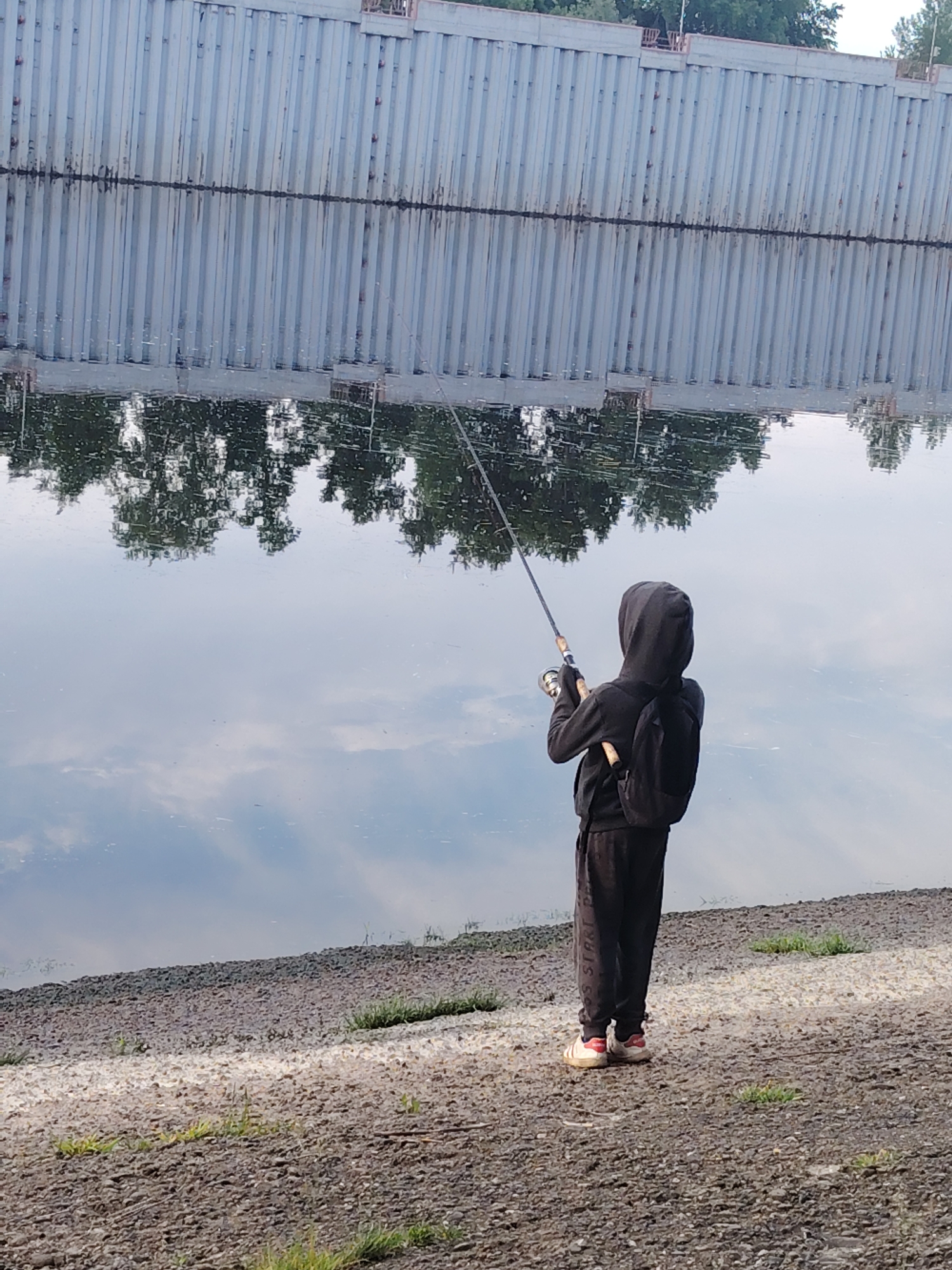 A person is seen from behind, fishing on the bank of a calm body of water, likely a river or canal, in Chaikovsky, Russia. The individual, dressed in a dark hooded sweatshirt, dark pants, and a dark backpack, holds a fishing rod angled out over the water. Their shoes are white with red accents and some faint, illegible text on the side, possibly "STARI". The pants also have some dark, indistinct text or a logo on the lower leg.
The water surface is smooth and highly reflective, mirroring the surroundings. Directly across from the fishing person is a tall, light-colored (possibly concrete or metal) retaining wall with distinct vertical ridges. The wall appears to extend quite high, suggesting a significant drop or embankment. A dark, irregular line, possibly rust or a waterline, runs horizontally along the wall at the water's surface. Above the wall, some distant trees and sky are visible.
The water's reflection also shows an upside-down image of lush green trees, suggesting they are on the opposite bank or behind the photographer. Further out on the water, reflections of a cloudy sky are visible. The bank where the person stands is a mix of small rocks and gravel, with scattered patches of green grass. The overall scene suggests a tranquil moment of leisure activity, likely during daytime, with clear weather indicated by the reflections of the sky.