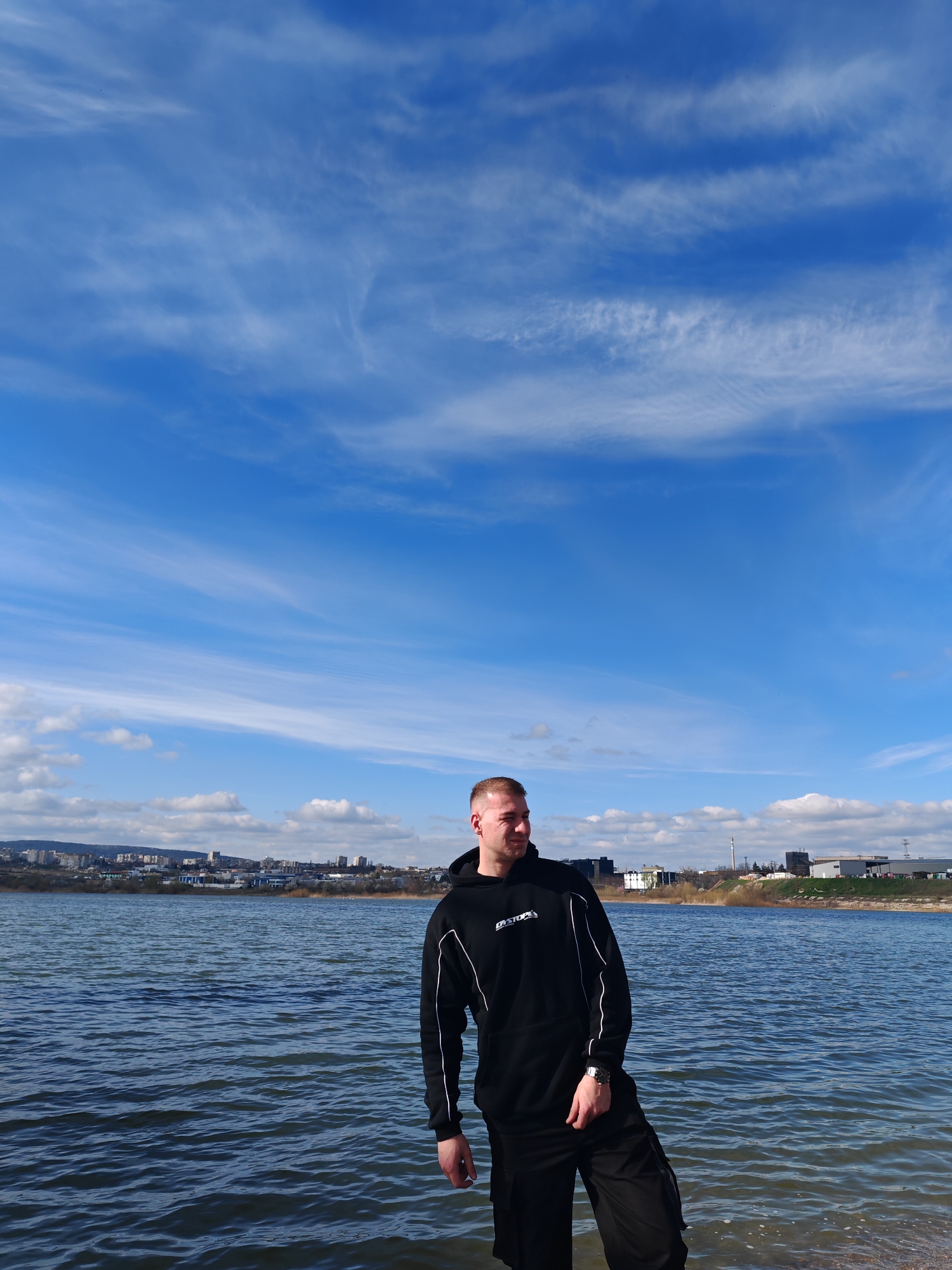 A medium shot shows a man standing near a body of water. He is fair-skinned with short blond hair and wearing a black hoodie and dark pants. His left hand rests in his pocket, and a silver watch adorns his left wrist. The man looks toward the left of the frame, away from the camera.
Behind him is a body of water with ripples on the surface. Across the water, a cityscape with low-rise buildings and industrial structures stretches across the horizon under a bright blue sky with wispy white clouds. The location is likely near the shores of Lake Studen Kladenets, in Shumen, Bulgaria. The terrain on the opposite shore suggests it is the industrial area of the city.