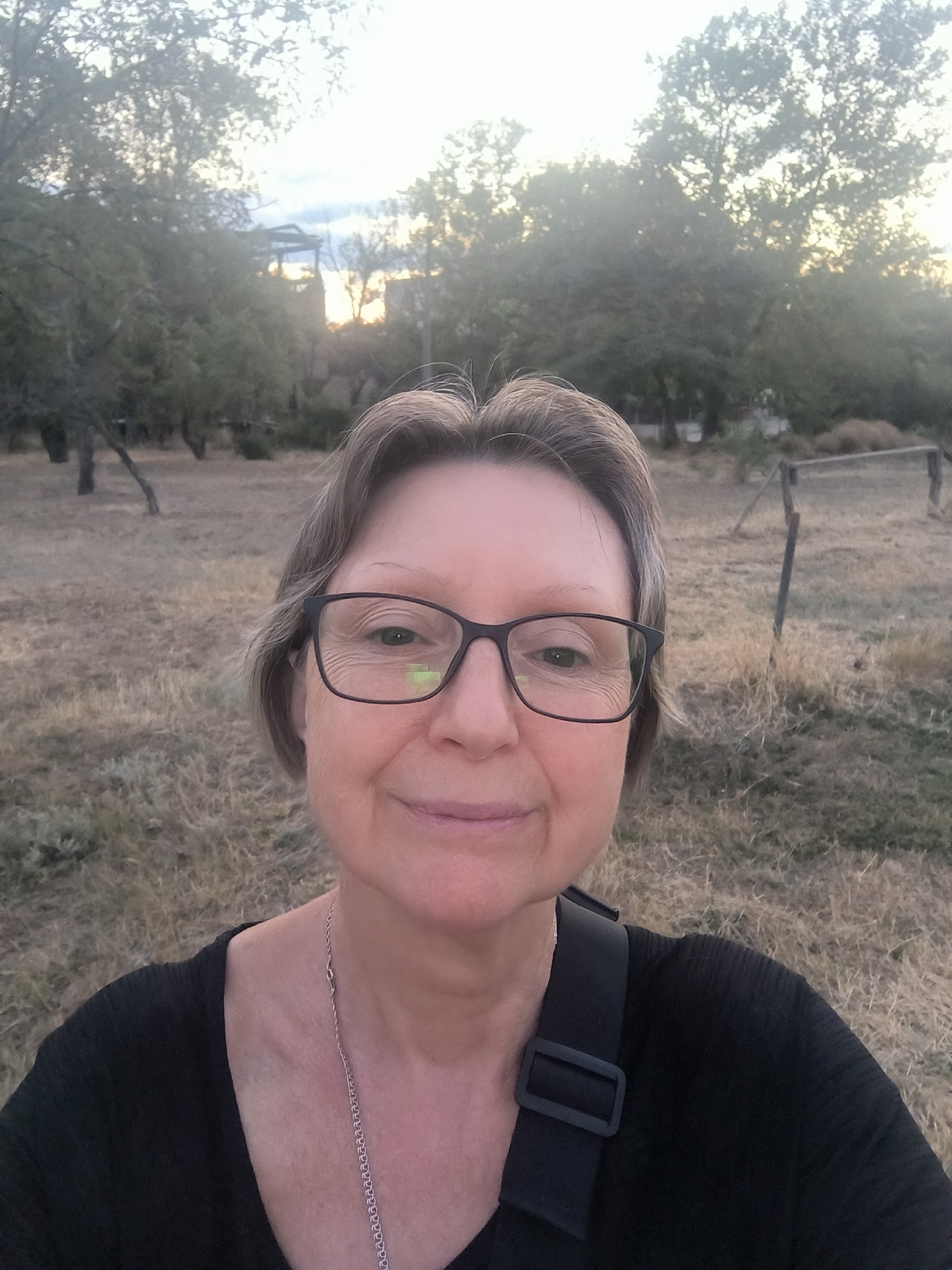 A woman is taking a selfie outdoors with a blurred background of trees and dry grass. She is wearing glasses and a black shirt. There is a strap from a bag visible across her shoulder. The environment appears to be a rural or park-like setting, with some structures visible in the distance. The lighting suggests it is either sunrise or sunset.