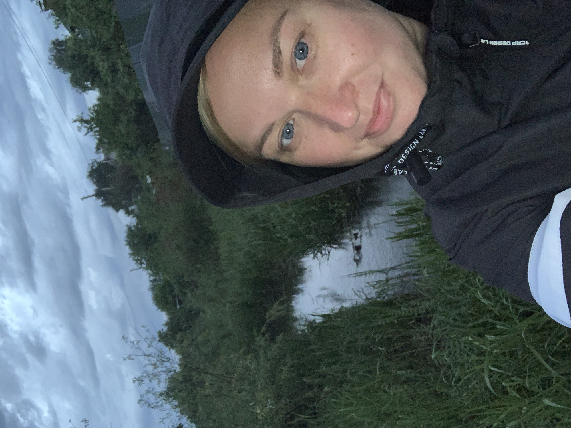 A woman with blonde hair and blue eyes wearing a black hooded jacket is taking a selfie. The hood is up. Behind her is a green, grassy area and a small body of water. The sky is overcast. Based on the user prompt, the location is Chaikovsky, Russia.