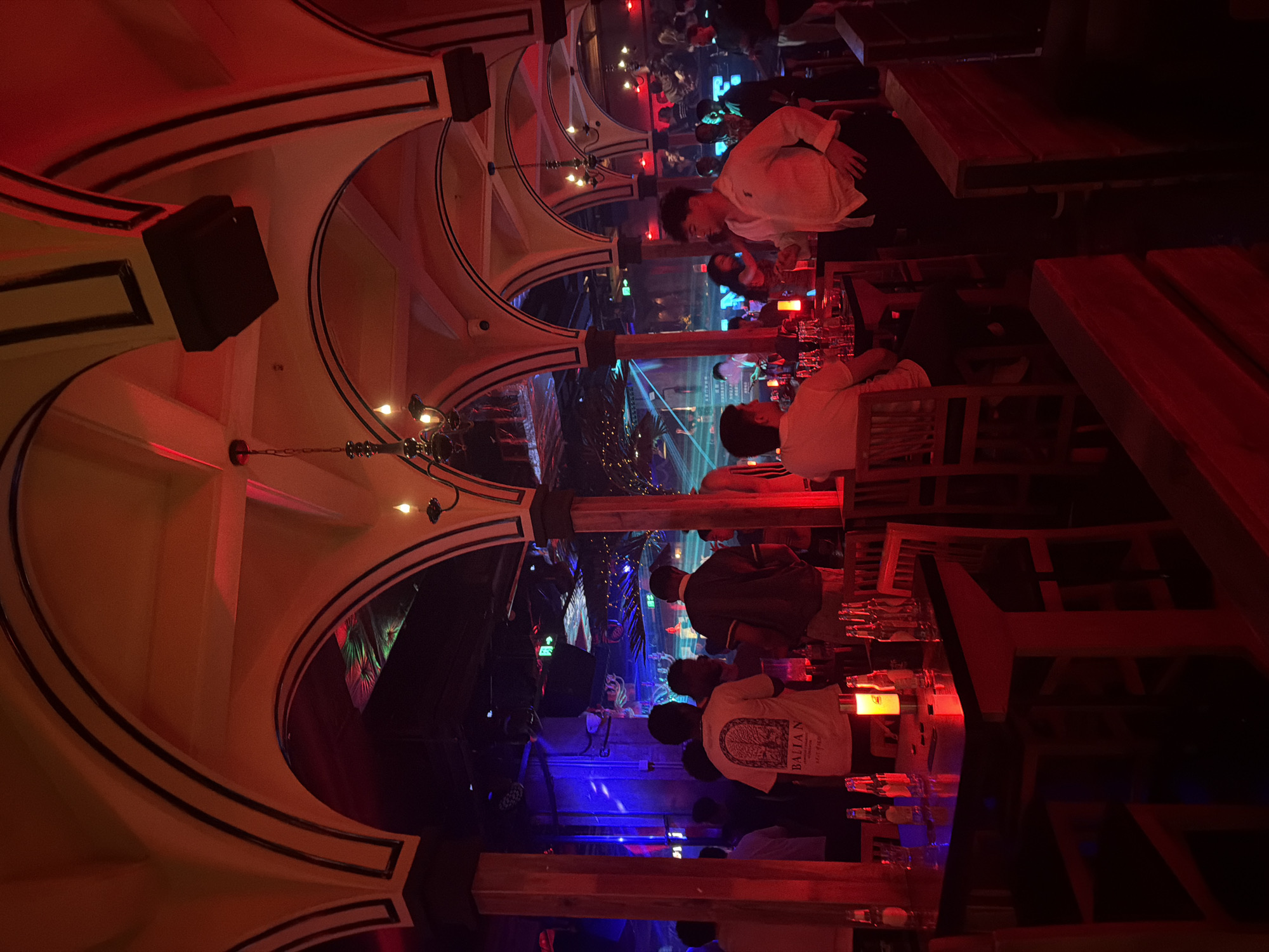 The image shows the interior of a dimly lit bar or club. The vaulted ceiling has decorative arches. Speakers are mounted high on the walls. People are seated at tables, and there are multiple glasses and bottles on the tables. The scene is lit with red and blue lighting, creating a nightclub atmosphere. The exact location in Xuanwu District, China, cannot be determined with certainty.