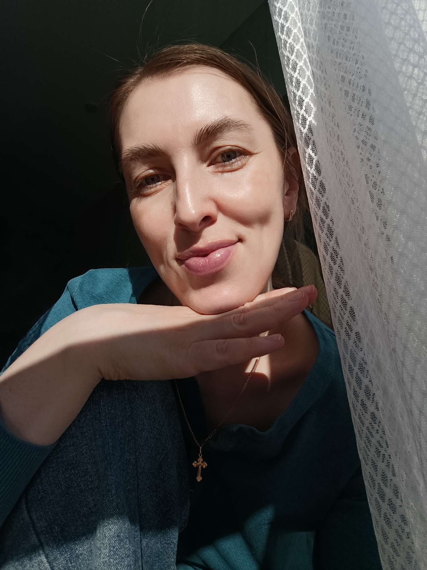 A woman poses in front of a window with a white lace curtain. She is wearing a teal shirt and denim jeans. A gold necklace with a cross pendant is visible. Sunlight illuminates her face and the curtain. The location is not discernible from the image.