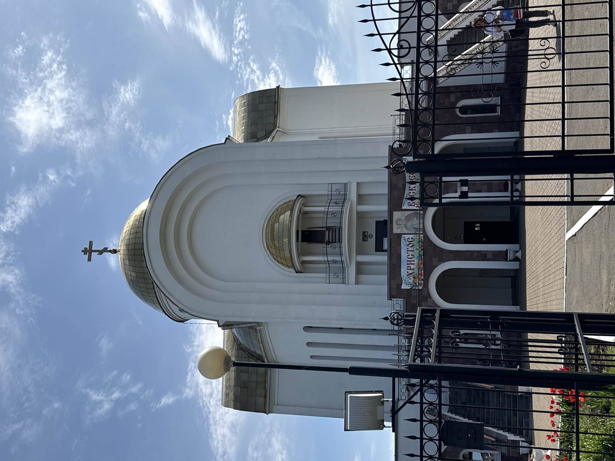 A ground-level, daytime shot shows the Dormition Cathedral in Votkinsk, Russia, viewed at an angle that depicts the church partially turned on its side. The building is white with a golden dome surmounted by a cross, set against a cloudy blue sky. The facade includes an arched opening with a balcony. A decorative sign is visible, written in Russian Cyrillic. In the foreground is a black metal fence. A lamppost and a boxy light fixture are also present. The setting appears to be a clear day with bright sunlight. The fence partially obscures the view of the church's entrance and some landscaping with red flowers.