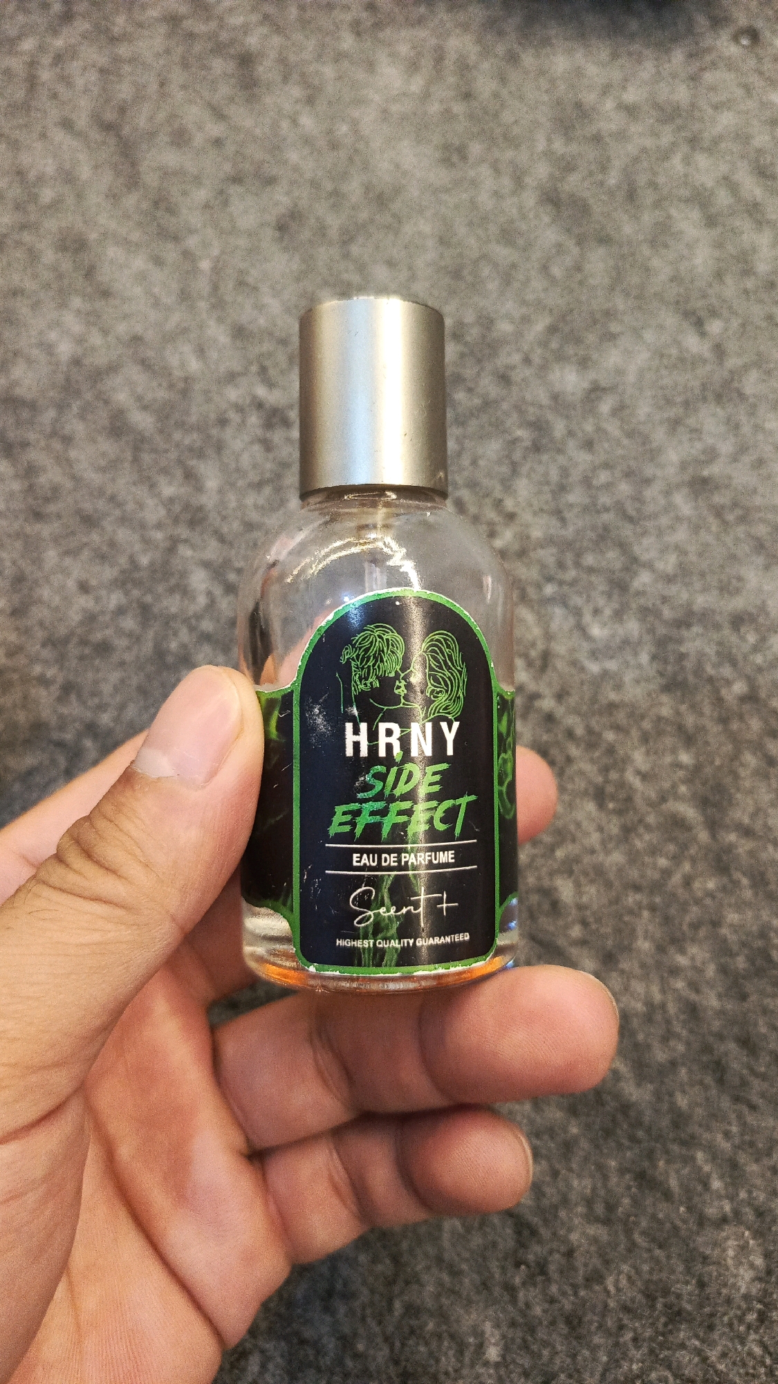 A hand is holding a small glass bottle of perfume. The bottle has a silver cap and a label with black and green text that reads "HRNY SIDE EFFECT EAU DE PARFUME Scent+". Below this, it says "HIGHEST QUALITY GUARANTEED". The background is a textured gray surface, possibly a carpet or fabric. The image was taken in Babadan, Indonesia.