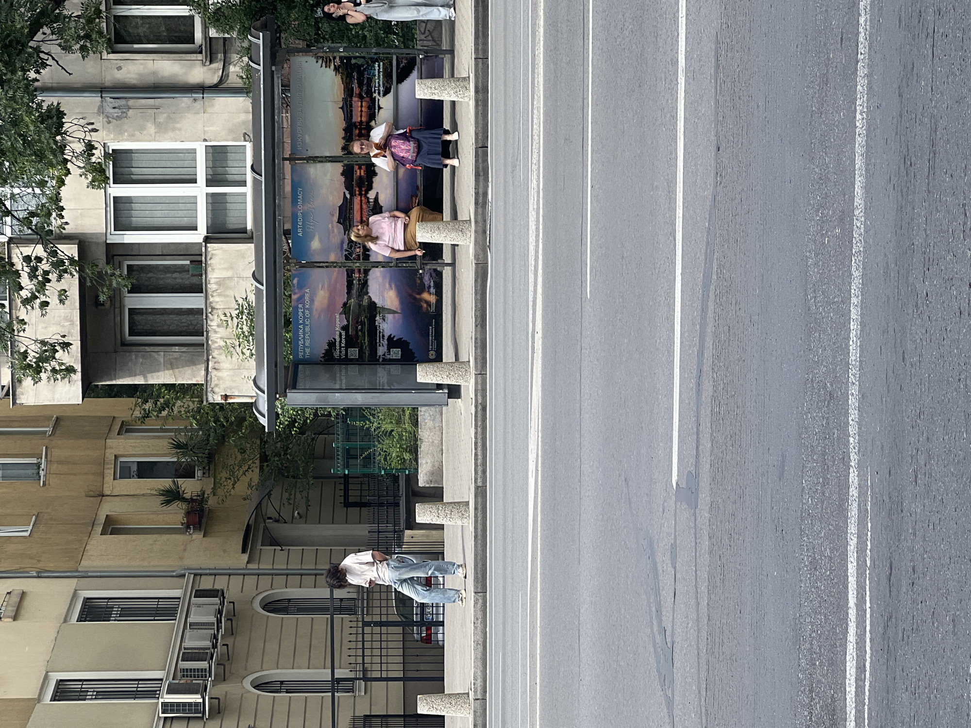 The image depicts a street scene in Sofia, Bulgaria, likely near a bus stop. In the foreground, a paved street with white lane markings is visible. A raised sidewalk runs along the street, bordered by concrete bollards. Behind the sidewalk is a bus stop shelter with several people waiting. One person, standing, is looking down at their phone, wearing a light-colored t-shirt and jeans. Two other individuals are seated on the bench inside the shelter. One, a woman with blonde hair, is wearing a pink shirt and brown skirt and appears to be smoking. The other, a woman with glasses, is wearing a white top and a dark skirt, and is looking at her phone. A third person, a woman in light-colored overalls, stands to the right of the bus stop. Behind the bus stop are buildings, one of which has air conditioning units mounted on its facade. Trees with green foliage are present, casting shadows. The overall impression is a typical urban environment on a bright, overcast day.