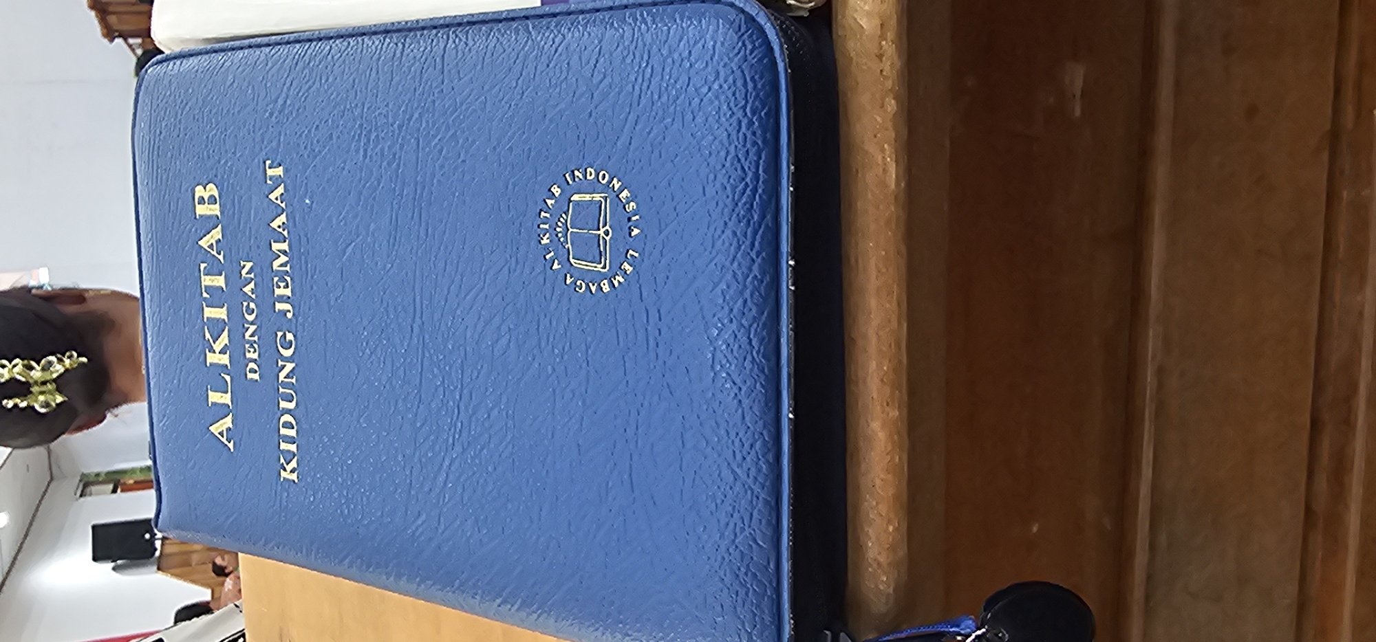 An open blue Bible with gold lettering is placed on a wooden surface. The Bible's cover features the text "ALKITAB DENGAN KIDUNG JEMAAT" in gold. In the lower right portion of the cover, a circular emblem with gold lettering and an open book design is embossed. A woman with her hair in an updo, adorned with a gold hair accessory, is partially visible in the background, facing away from the camera. The environment appears to be indoors, possibly a place of worship or meeting, suggested by the wooden furniture and the background elements. No specific location in Puncak Jaya, Indonesia, can be identified from the image.