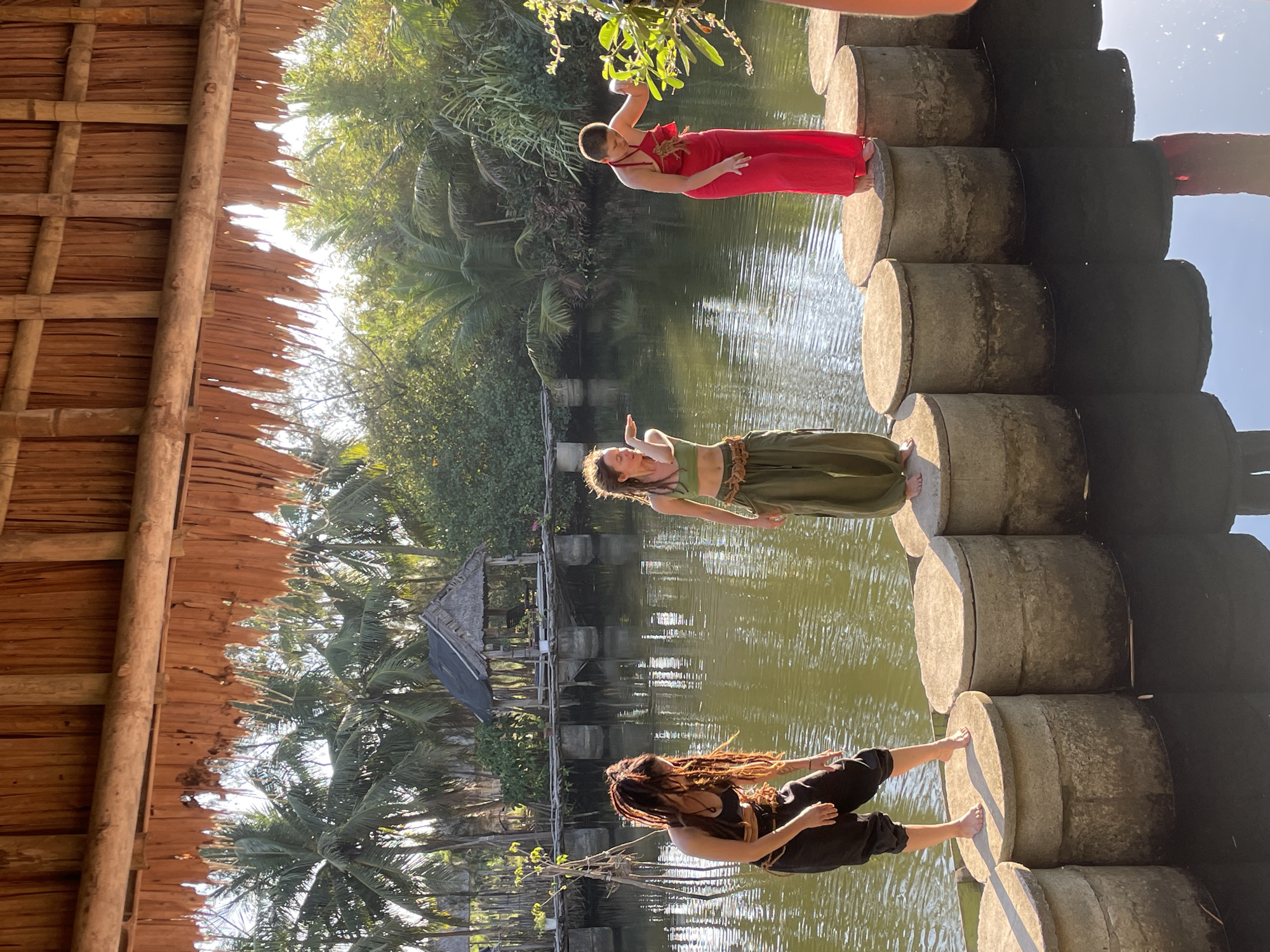 Three individuals are barefoot and stand on a series of concrete cylinders placed in a body of water that reflects the sky and surrounding vegetation. One individual, located at the top of the frame, wears a red dress. The individual in the middle is wearing green clothing. The third person at the bottom of the frame is wearing dark clothing and has long dreadlocks. The background consists of a variety of lush green trees and a thatched-roof structure partially visible in the top left corner of the frame. A small building is seen beyond the water. The location may be Đà Nẵng, Vietnam.