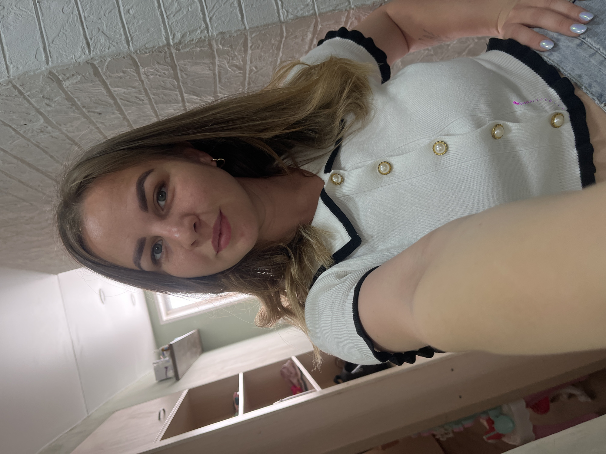 A woman is taking a selfie. She is wearing a white shirt with black trim and pearl buttons, and light-wash denim shorts. She has shoulder-length blonde hair and is wearing gold heart-shaped earrings. The background shows the interior of a room with a white brick-patterned ceiling, white cabinets, a window, and a sink. The location may be in Izhevsk, Russia, but without further information, this cannot be confirmed.