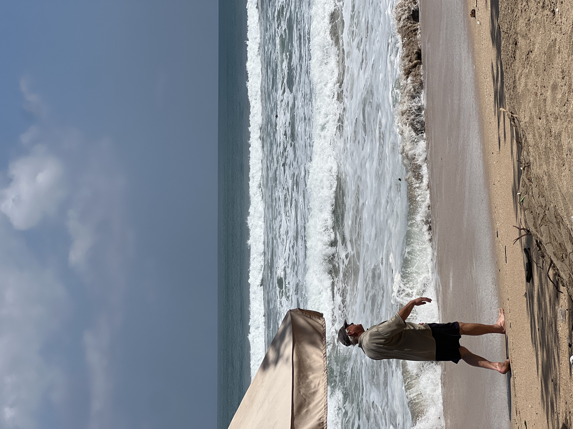 An older man wearing a light-colored short-sleeved shirt, dark shorts, and a baseball cap is walking barefoot on a sandy beach in Kuta, Indonesia. He is walking away from the camera, towards the ocean. The man is partially shaded by a large, light-colored beach umbrella. The ocean has moderate waves with whitecaps, and the sky is overcast with some patches of lighter sky. The beach is composed of light brown sand, with some debris and driftwood scattered on it.
