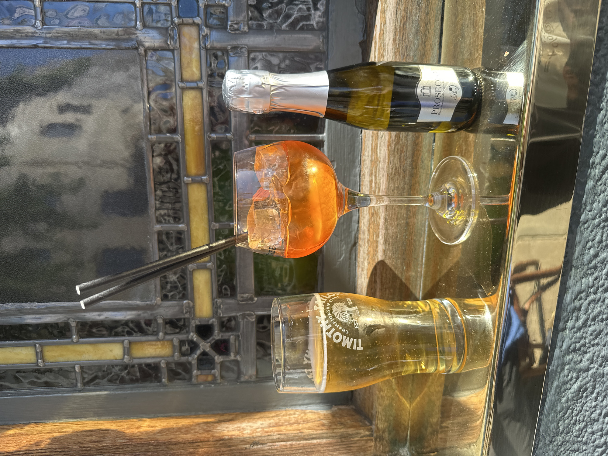 A still life scene features a glass of beer, a cocktail, and a small bottle of Prosecco arranged on a wooden surface in front of a stained-glass window. The beer is in a pint glass with the "Timothy Taylor" logo visible. The cocktail, an orange aperitif with ice, is in a wine glass. The Prosecco bottle is small with a silver foil cap and a label that reads "Prosecco Denominazione di Origine Controllata." Two black straws are placed in the cocktail glass. The stained-glass window has a pattern of yellow and darker colored panes. The overall setting suggests an outdoor seating area, possibly a pub or restaurant, given the drinks and the decorative window. No people are present in the image. The lighting indicates it is daytime, with sunlight casting shadows.