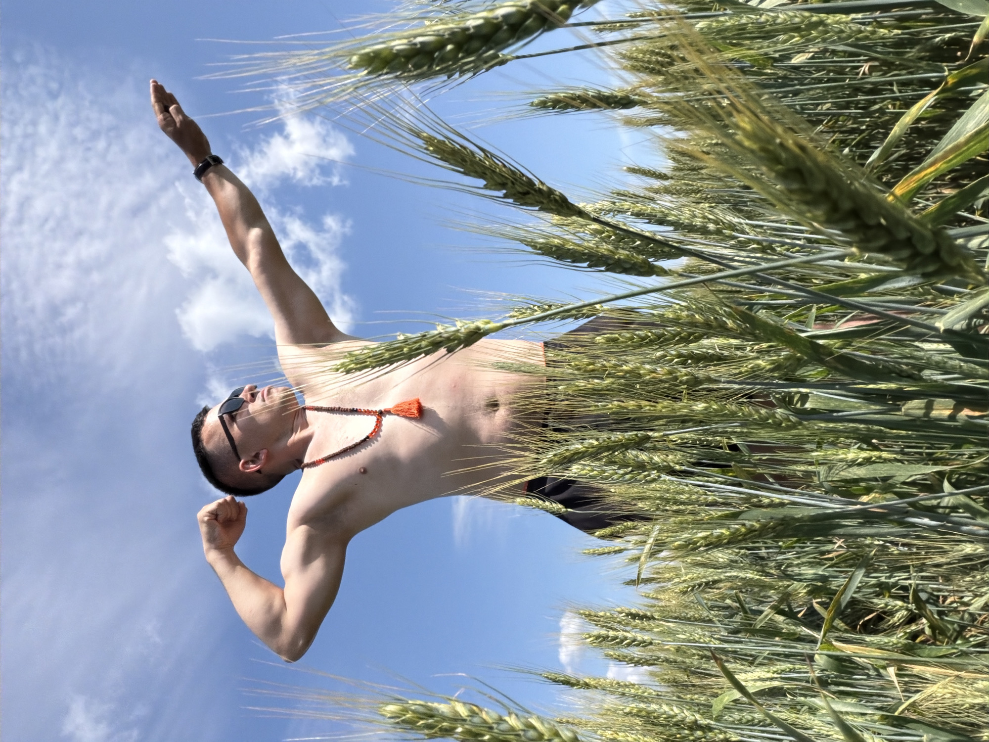 A shirtless man wearing sunglasses and a beaded necklace with an orange tassel poses in a wheat field under a blue sky with scattered clouds. His left arm is flexed at the elbow, and his right arm is extended horizontally to his right, with his hand in a flat, open position. The man appears to be standing in a field of mature wheat, with the stalks of wheat prominent in the foreground and surrounding him. The sky is bright and clear, indicating daytime. The location in Podshivalovo, Russia is not discernible from the image itself.