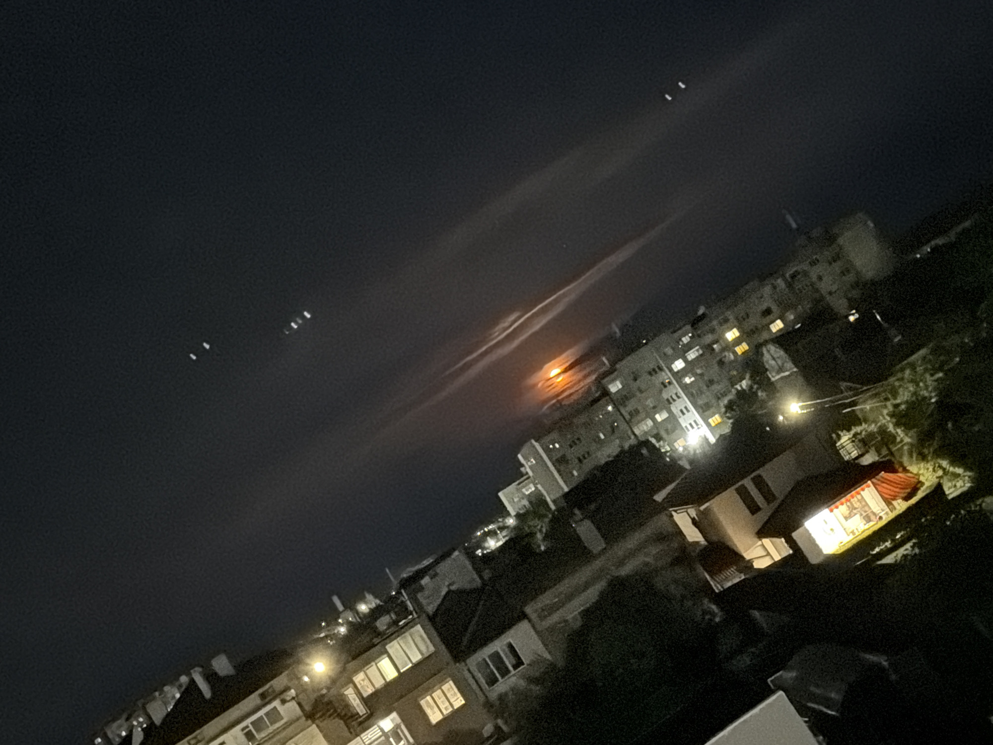 The image shows a nighttime urban scene, likely in Burgas, Bulgaria. The view is from an elevated position, overlooking residential buildings and the city skyline. The sky is dark, with a thin layer of clouds and a bright orange object shining through, probably the moon. Several buildings are illuminated with interior lights, and a distinct house with a red roof and bright interior light is visible in the foreground. Three distinct lights are noticeable in the upper left corner of the image, suggesting either celestial objects or artificial lights. The scene appears calm and quiet, with no visible people or clear interactions.
