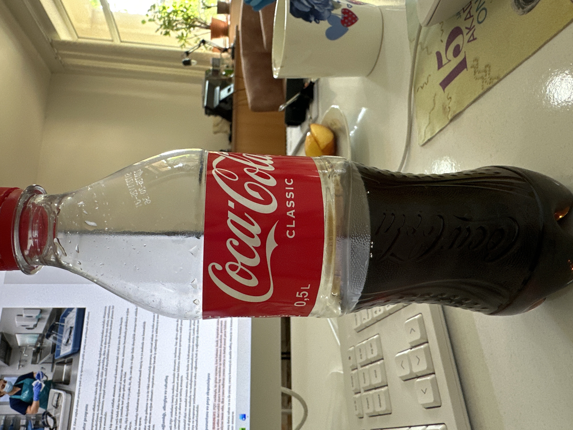 The image shows a Coca-Cola Classic 0.5L bottle on a white desk. The bottle is partially filled with dark brown liquid and condensation droplets are visible on the outside. To the left of the bottle is a white keyboard. In the background, a document is visible with text and an image of a person wearing gloves and a mask in a laboratory setting. To the right of the bottle, there is a white mug with a floral design and two heart shapes, and a small plate with orange wedges. Further in the background, a window with a view of plants is visible, as well as office furniture. The overall scene appears to be an indoor office or workspace. There is no specific indication of the location being in the city of Baku, Azerbaijan.