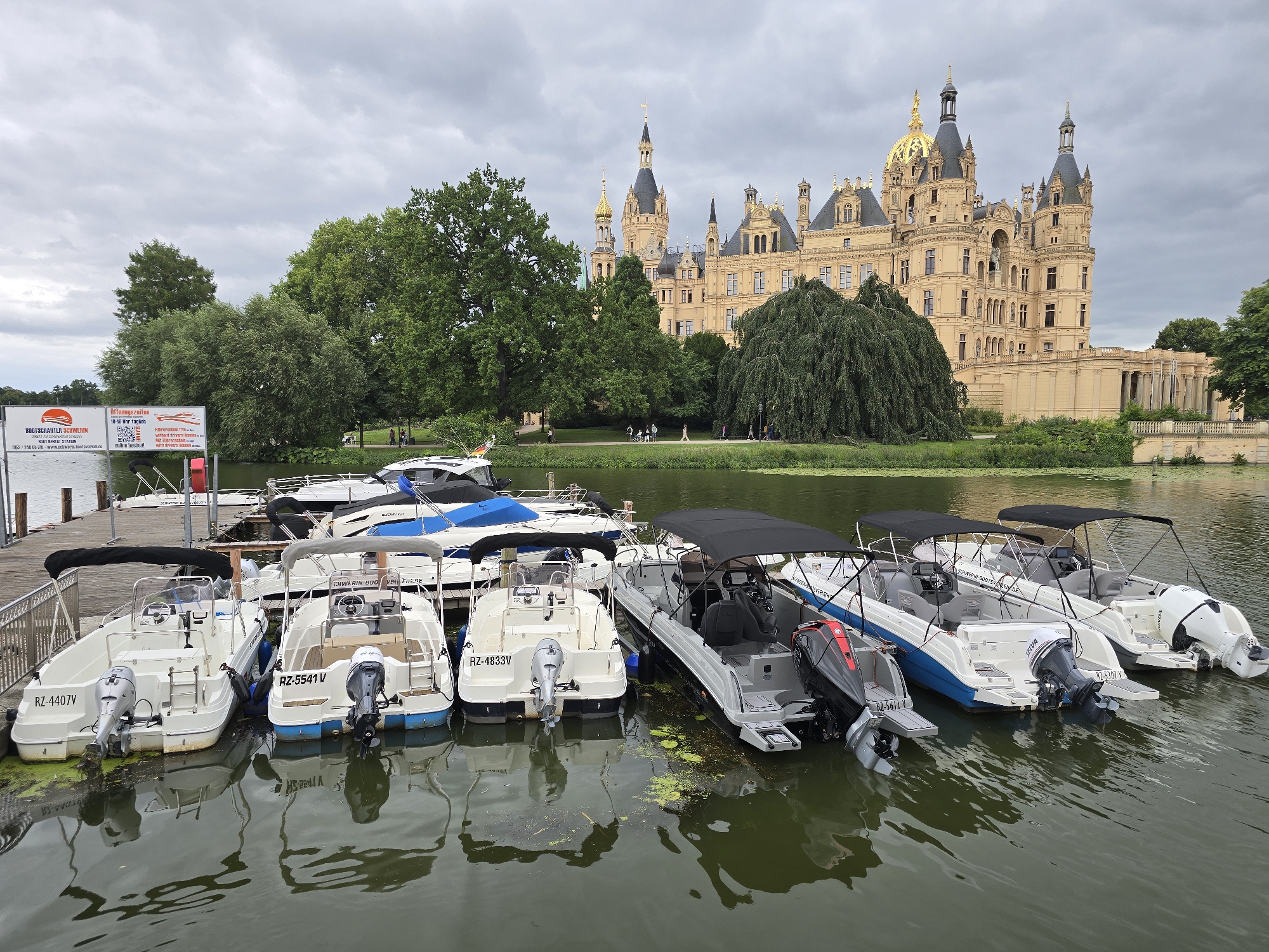The image depicts a fleet of small white motorboats docked at a wooden pier on a cloudy day. In the background, Schwerin Castle, a prominent landmark in Schwerin, Germany, stands tall with its distinctive spires and golden domes. The castle is situated on an island surrounded by lush green trees and vegetation. The boats are arranged in multiple rows, with their reflections visible in the calm water. Some of the boats have outboard motors, while others are equipped with covered canopies. The pier is accompanied by a sign advertising boat rentals. A few people are visible walking along the pathway in front of the castle. The overall scene is tranquil and picturesque, showcasing a harmonious blend of nature and architecture.