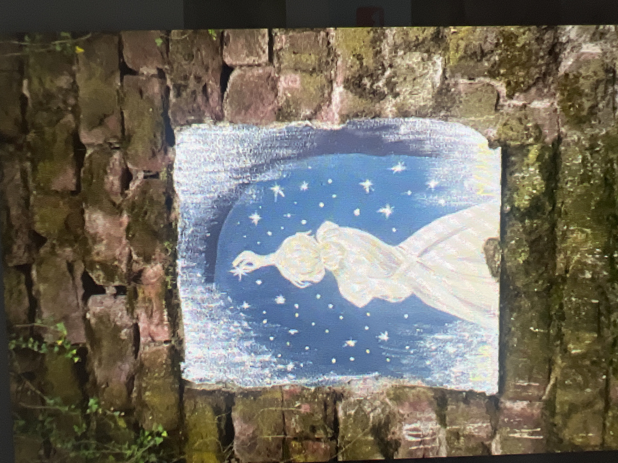 A rectangular painted mural is embedded in a brick wall, presumably outdoors. The brick wall appears aged, with moss growth on some bricks. The mural features a figure, possibly an angel or a person in a gown, set against a blue background filled with stars. The figure is in a reclining or flying position, with an arm extended toward a star. The style is whimsical. The mural is located in the city of Sofia, Bulgaria.
