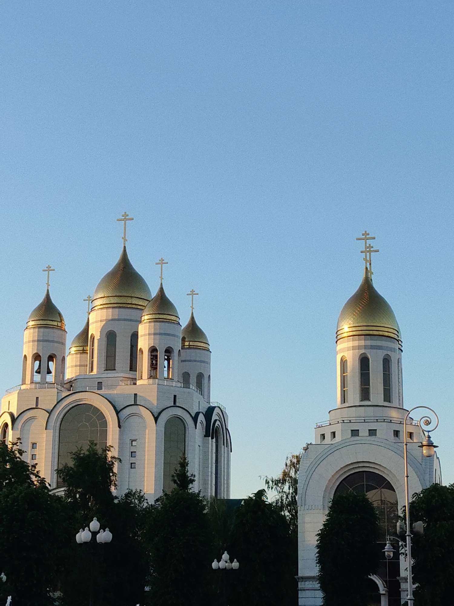 The image shows a large, white cathedral with golden domes against a clear blue sky. The cathedral has multiple towers, each topped with a cross. The main building is intricate, with arched windows and decorative detailing. In the foreground, there are dark green trees and several ornate street lamps. The lighting suggests it is either early morning or late afternoon, as the sun casts a warm glow on the cathedral's facade. The scene is peaceful and architectural.