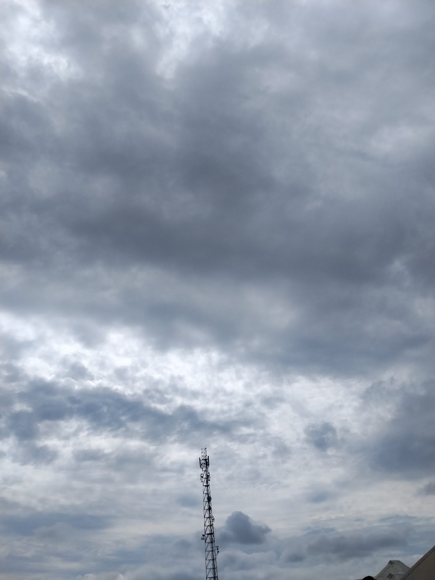 The image shows a view looking up at a cloudy sky with a cell tower on the right side. The sky is filled with dark gray and white clouds, suggesting overcast weather. The cell tower is tall and slender, with various antennas and equipment attached to it. The lower portion of the image shows a sliver of rooftops, indicating an urban or semi-urban environment. The scene is likely captured in Mangu Town, Nigeria, given the context.