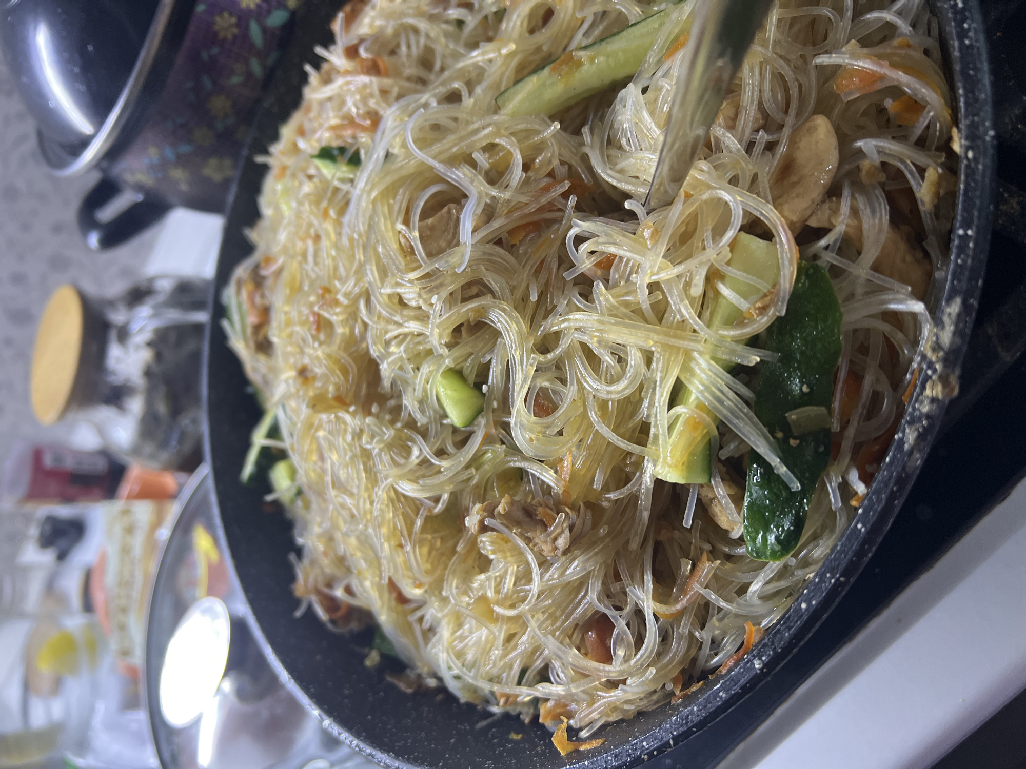 The image shows a close-up of a dark-colored pan filled with a dish of stir-fried glass noodles. Mixed with the noodles are various ingredients, including sliced cucumbers, shredded carrots, and pieces of chicken or tofu. A pair of metal tongs is lifting a portion of the noodles. The pan sits on a white surface, likely a stovetop, with other kitchen items visible in the background, including a pot with a floral pattern and a clear glass bowl. The location and city of Noviy, Russia, cannot be determined from the given image.