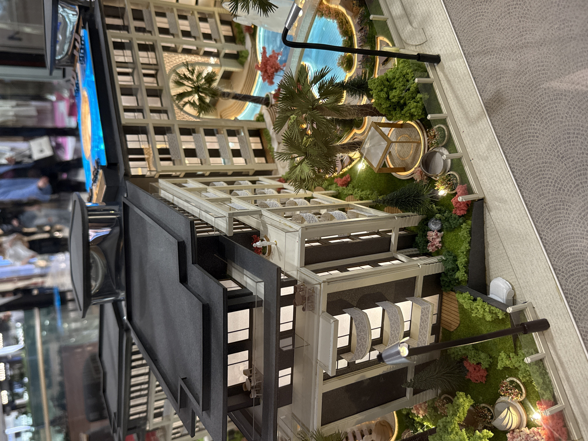 The image depicts a miniature model of a building complex. The model features multiple structures, including a tall high-rise building, possibly a residential or commercial space. In addition, a lower structure with gardens, palm trees and what appears to be a pool is also visible. The model also depicts street lamps and other decorative elements, presumably to illuminate it. The scale model sits on a patterned base, maybe mimicking pavement. The lighting and general detailing suggest it is a professional architectural model, potentially for marketing. The location is not evident in the image, it is just a model.
