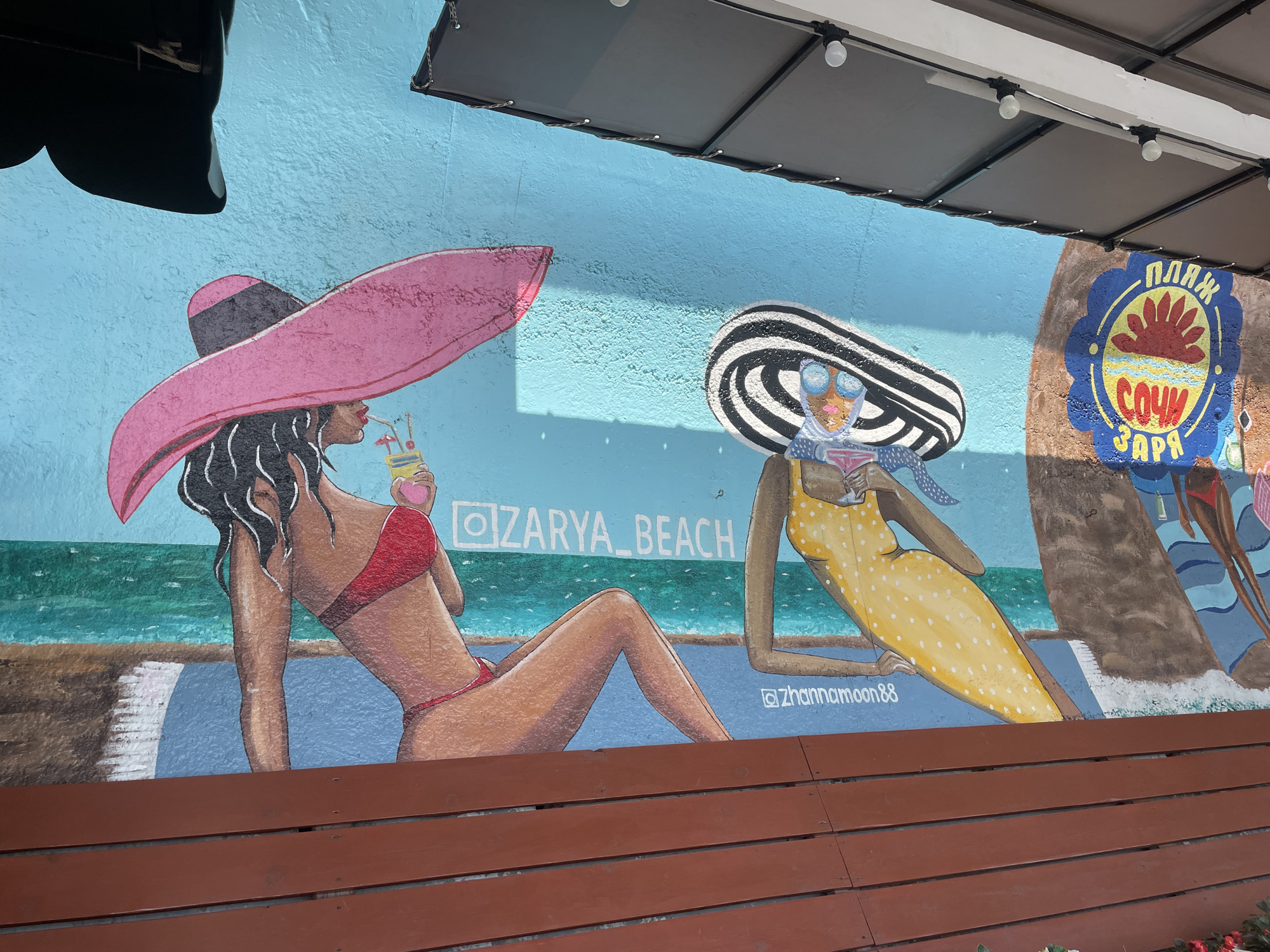 The image shows a mural painted on a blue wall, depicting a beach scene in Sochi, Russia. Two women are the main subjects. The woman on the left is wearing a red bikini and a large pink sunhat. She is holding a drink with a straw and a piece of fruit. The woman on the right is wearing a yellow polka-dot dress and a striped sunhat. She has sunglasses on and is holding a drink. The background of the mural features a turquoise ocean and a sandy beach. To the right of the women, there is a circular emblem with text in Russian that reads "ПЛЯЖ СОЧИ ЗАРЯ," which translates to "Sochi Zarya Beach." The mural is located outdoors, as evidenced by the presence of a roof structure with lighting fixtures extending over the top of the mural. In front of the mural is a wooden bench. The overall scene is vibrant and colorful, evoking a summer vacation atmosphere.