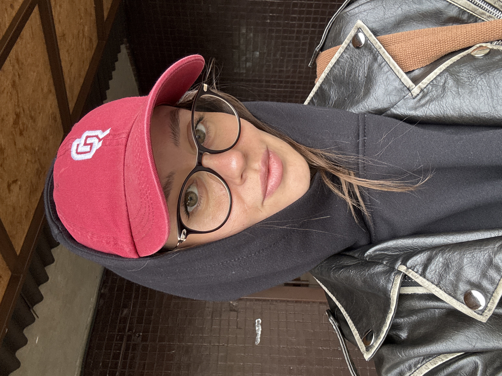 A woman wearing a red baseball cap with a white logo, round glasses, and a black hoodie is posing for a selfie. She is wearing a black leather jacket with silver accents. The background features a brown tiled surface, possibly a wall or floor. The photo was likely taken indoors or in a covered area. There is no information that indicates this photo was taken in Krasnoyarsk, Russia.