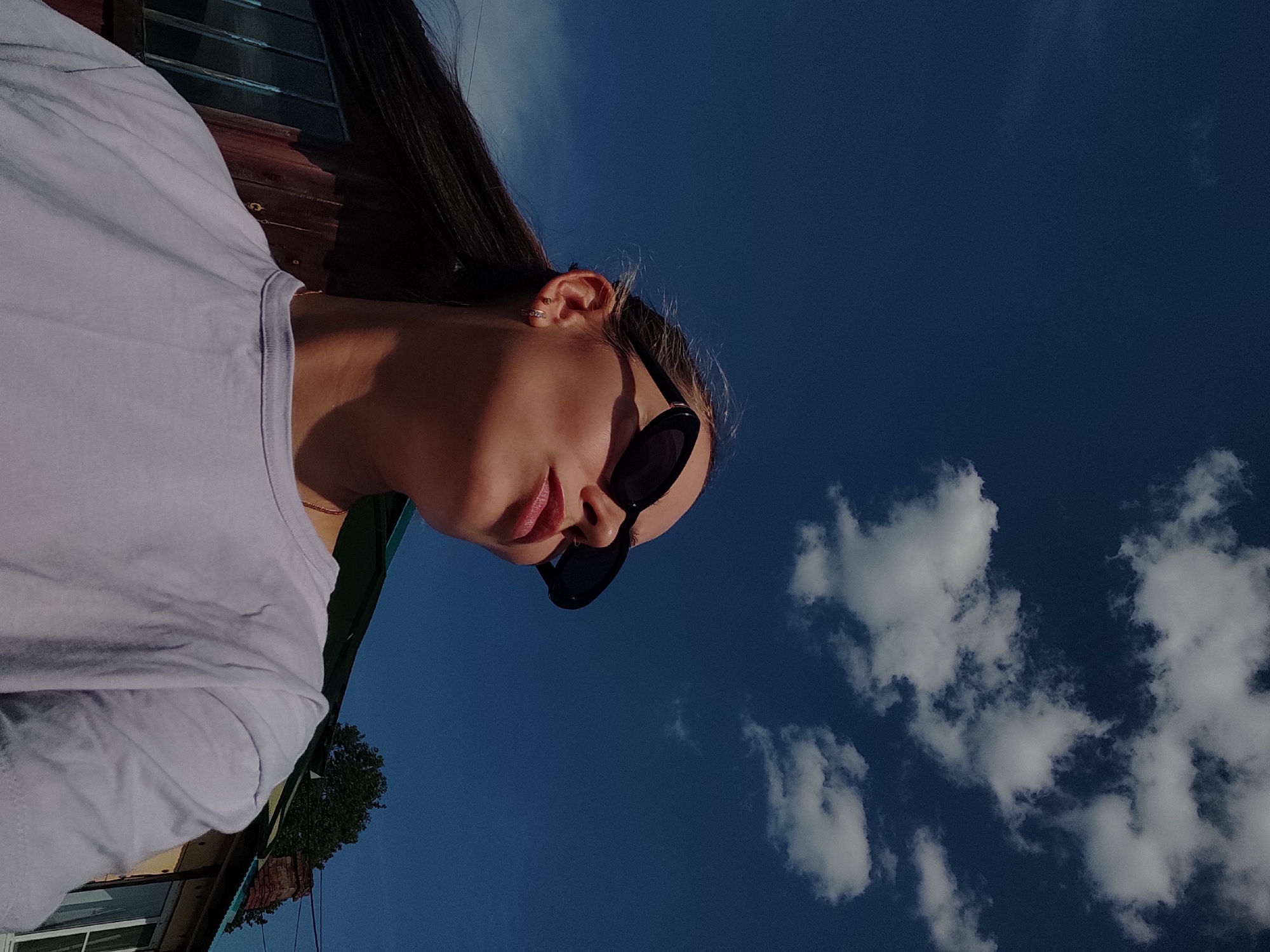 A low-angle, tilted photograph captures a woman outdoors in Ольховка, Russia. She is wearing black sunglasses and a light-colored t-shirt. The background consists of a clear blue sky with scattered white clouds. Some building structure elements, possibly parts of a building or porch, are visible in the periphery of the frame. A tree is partially visible. The light suggests it is a sunny day.