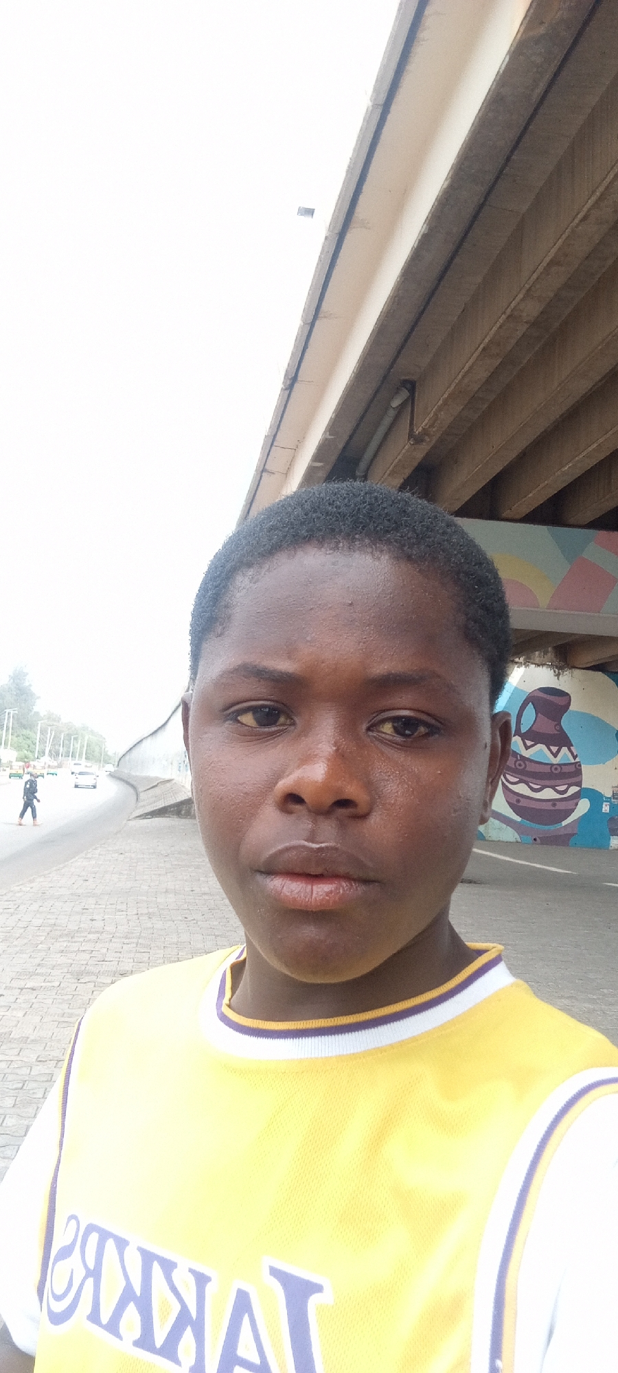 The image is a medium close-up selfie of a young person, appearing male, with short, dark, closely-cropped hair and a neutral expression, looking directly at the viewer. Their face appears slightly shiny, suggesting warmth or humidity. The person is wearing a yellow athletic jersey with white short sleeves and purple and white striped trim around the collar and sleeve cuffs. The word "LAKERS" is visible in purple text across the chest, appearing reversed, indicating a front-facing camera shot.
The setting is an outdoor urban environment, likely in Jos, Nigeria, under or adjacent to a large concrete overpass or bridge. The underside of the bridge structure is visible in the upper right, showing support beams and a section of a colorful mural depicting a stylized pot or vase design in blues, purples, and browns.
To the left of the subject, a textured, paved sidewalk leads to a curved road with light traffic in the distance. A pedestrian is visible walking on the road further back, and several cars are present. The sky is bright and appears overcast or heavily exposed, creating a soft, diffused light. The overall scene captures a candid moment in a bustling city environment.