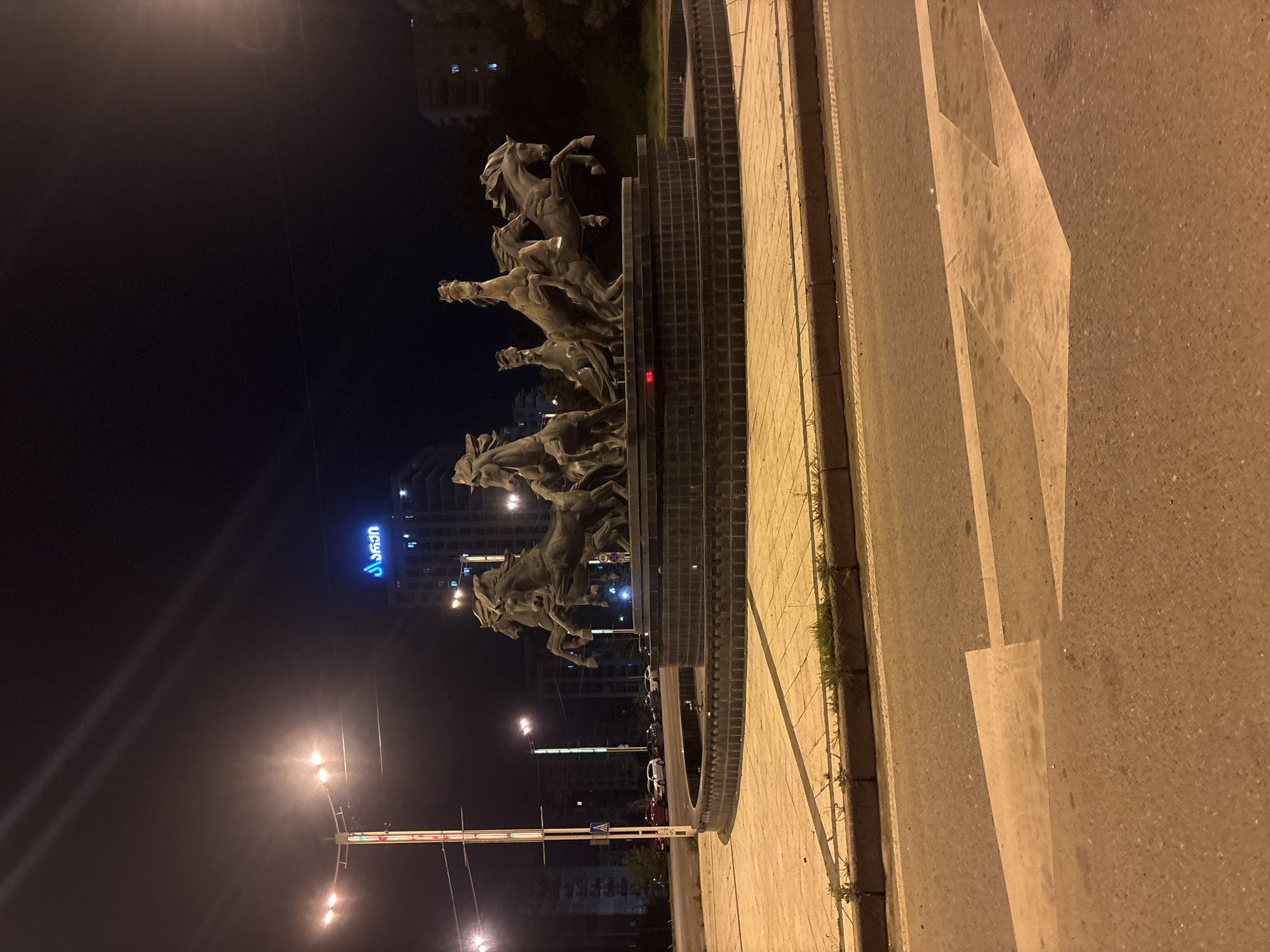 The image depicts the "Monument of King Vakhtang Gorgasali" in Tbilisi, Georgia, at night. The sculpture features a group of horses rearing up. The monument is situated near a crosswalk on a paved street, with a modern building lit with neon lights in the background. Streetlights are also visible in the foreground. The image is oriented sideways.