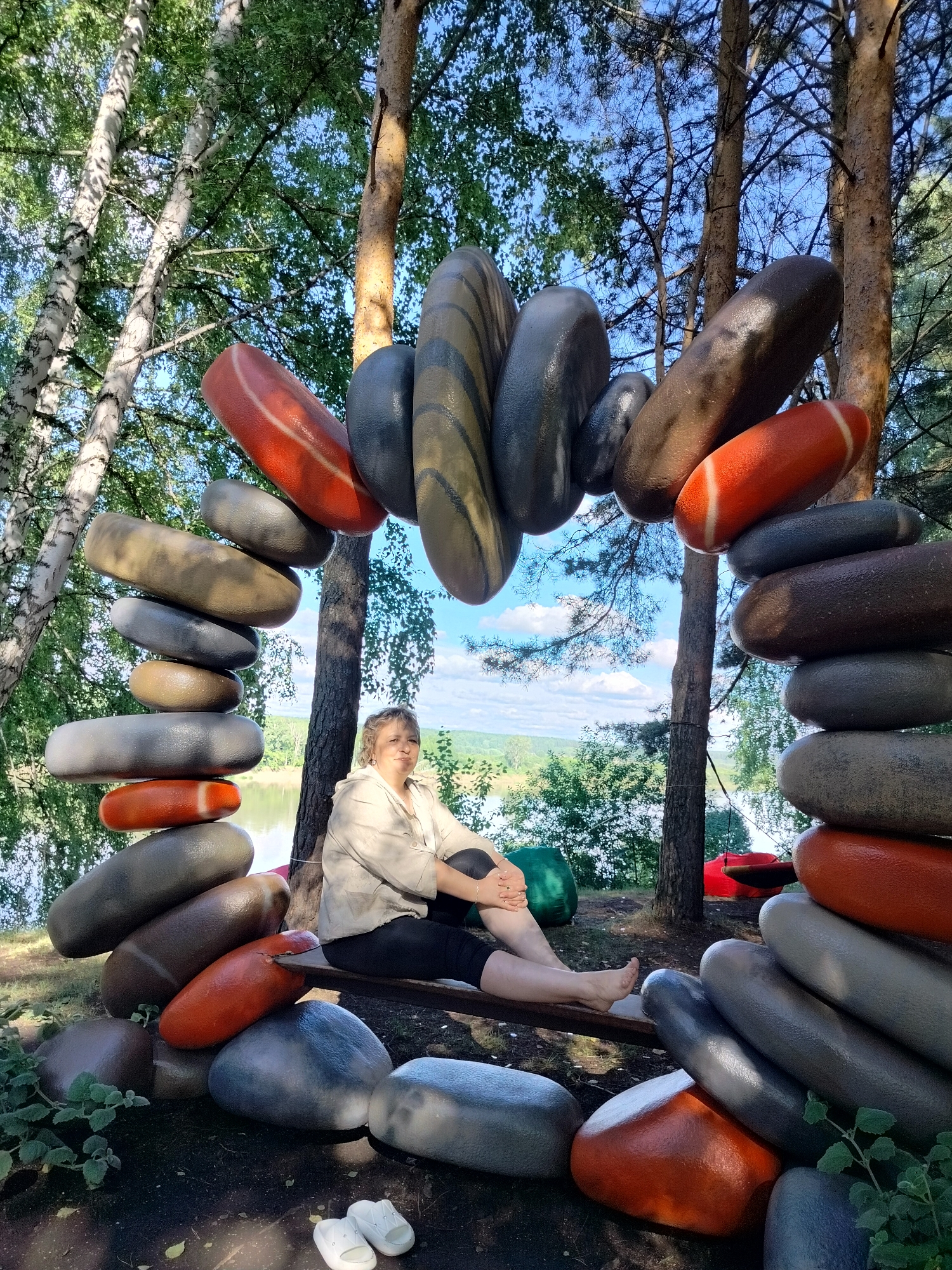 The image depicts a woman sitting on a bench outdoors. She is wearing a light-colored, loose-fitting top and dark pants. She is seated with her legs crossed and her arms resting on her legs. The woman appears to be in her late 50s or early 60s.
Behind and around the woman is a sculpture made of large, smooth, oval-shaped objects stacked in an arch and in piles. These objects are colored in shades of gray, brown, and orange, with some having striped patterns. The sculpture is placed in a wooded area with tall trees and greenery. In the background, a body of water, likely a lake or river, is visible, along with a partly cloudy blue sky. The ground is covered with dark soil and some fallen leaves. A pair of white flip-flops is on the ground in the foreground. Two large, beanbag-like chairs, one green and one red, are visible in the background among the trees. The scene is consistent with an outdoor recreational area in the village of Berezovo (selo), Novokuznetsky District, Russia, possibly near a body of water.