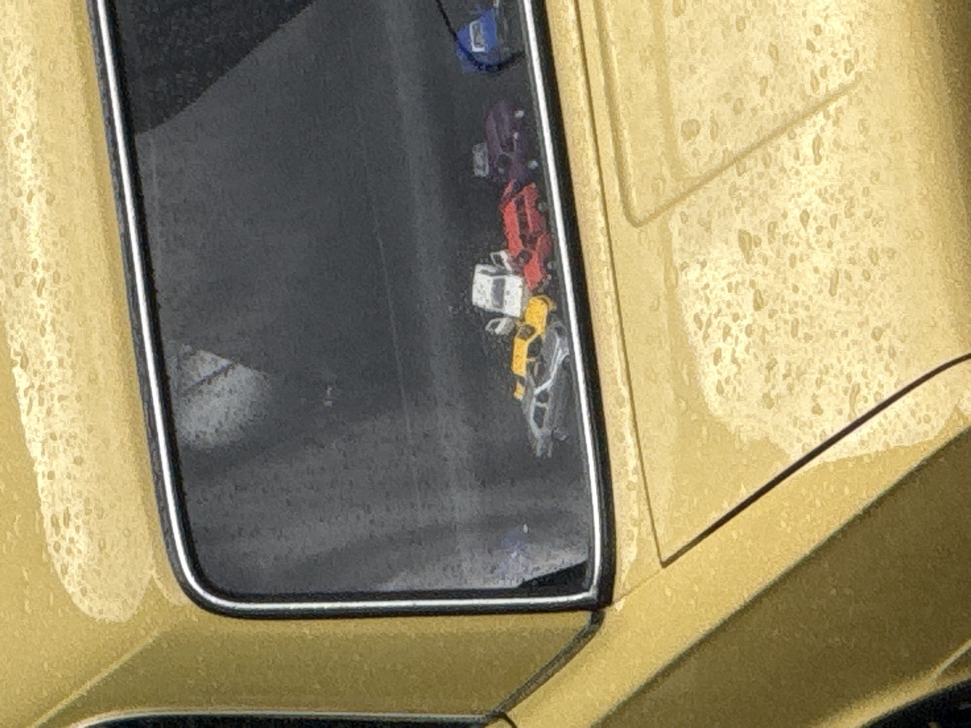 The image shows a close-up, eye-level shot of the rear window of a yellow car. The window is dirty and speckled with water droplets, suggesting it is raining or has recently rained. Inside the car, visible through the rear window, are several small toy cars. From left to right, there appears to be a silver car, a yellow car, a white car with its doors open, a red car, a dark purple car, and a blue car. The background outside the car is dark and out of focus. The setting is not identifiable as Moscow, Russia.
