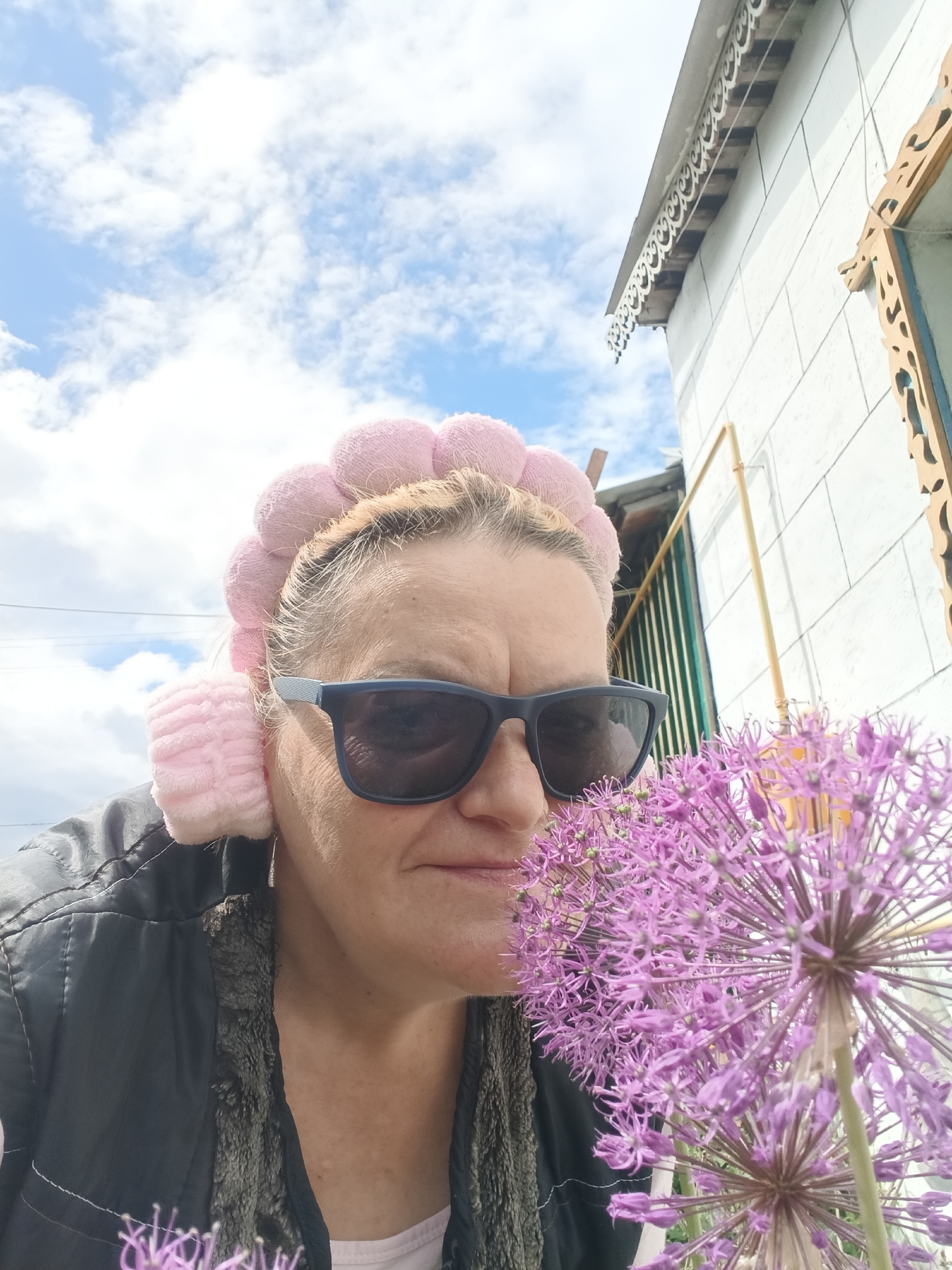 The photograph depicts a woman wearing pink hair rollers and dark sunglasses outdoors in Красный Маяк, Russia. She is positioned close to the camera, with a large, spherical purple flower partially obscuring the lower right of the frame. The woman is wearing a black vest over a light-colored shirt. Behind her, a white building with decorative wooden trim is visible, along with a glimpse of a blue and white sky with scattered clouds. A yellow pipe runs along the wall of the building. The scene is lit by natural daylight.