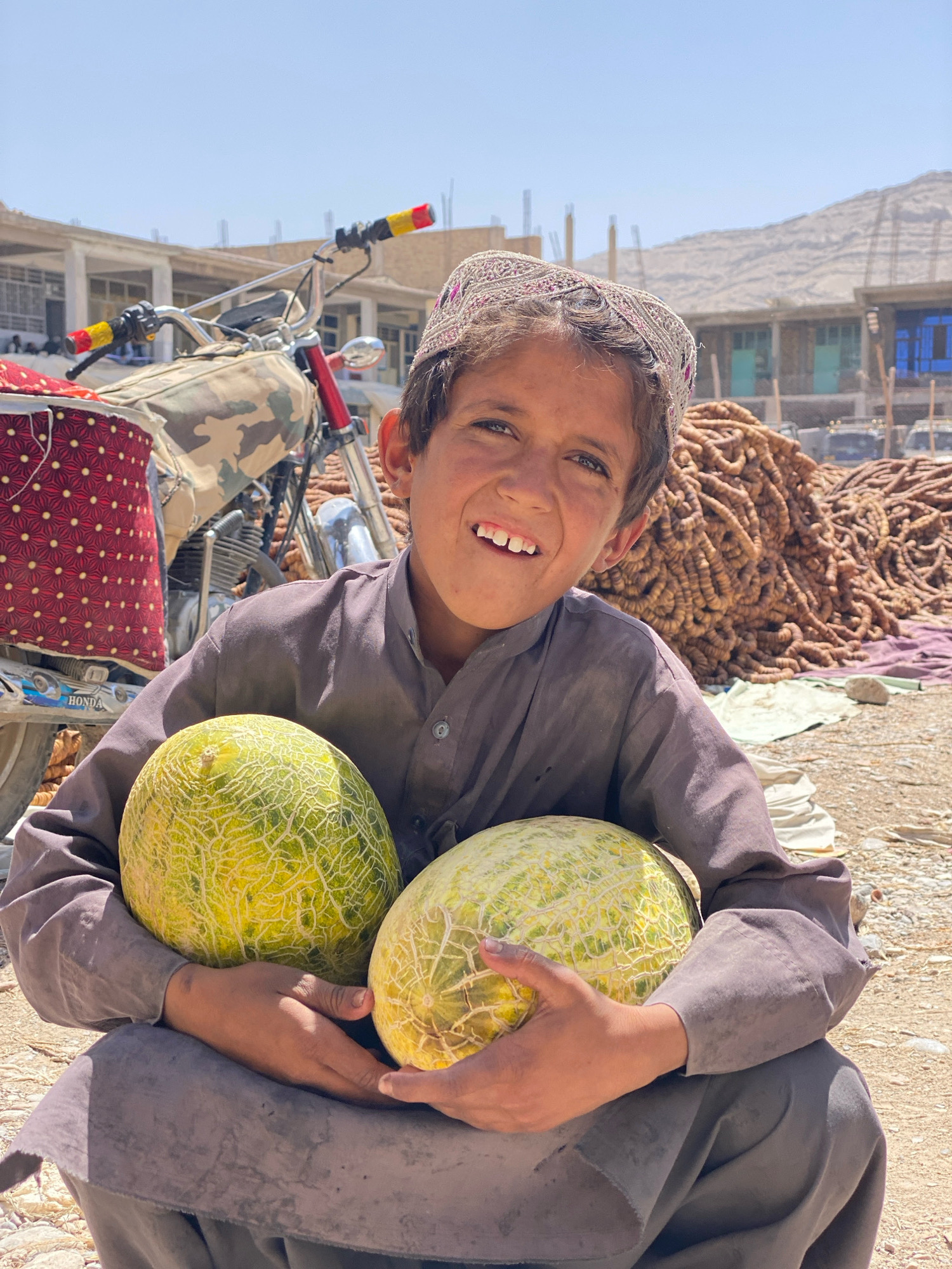 A young boy wearing a traditional embroidered cap and a dark grey shalwar kameez squats on dusty ground, holding two large, pale green melons. He has a wide smile, revealing a gap in his front teeth. Behind him, a camouflage-covered motorcycle is parked. Further in the background, a large pile of dried, twisted produce, possibly dates or mulberries, is visible. Beyond that, buildings with light-colored walls and blue-framed windows stand under a clear, bright sky. The scene appears to be an outdoor market in Kandahar, Afghanistan.