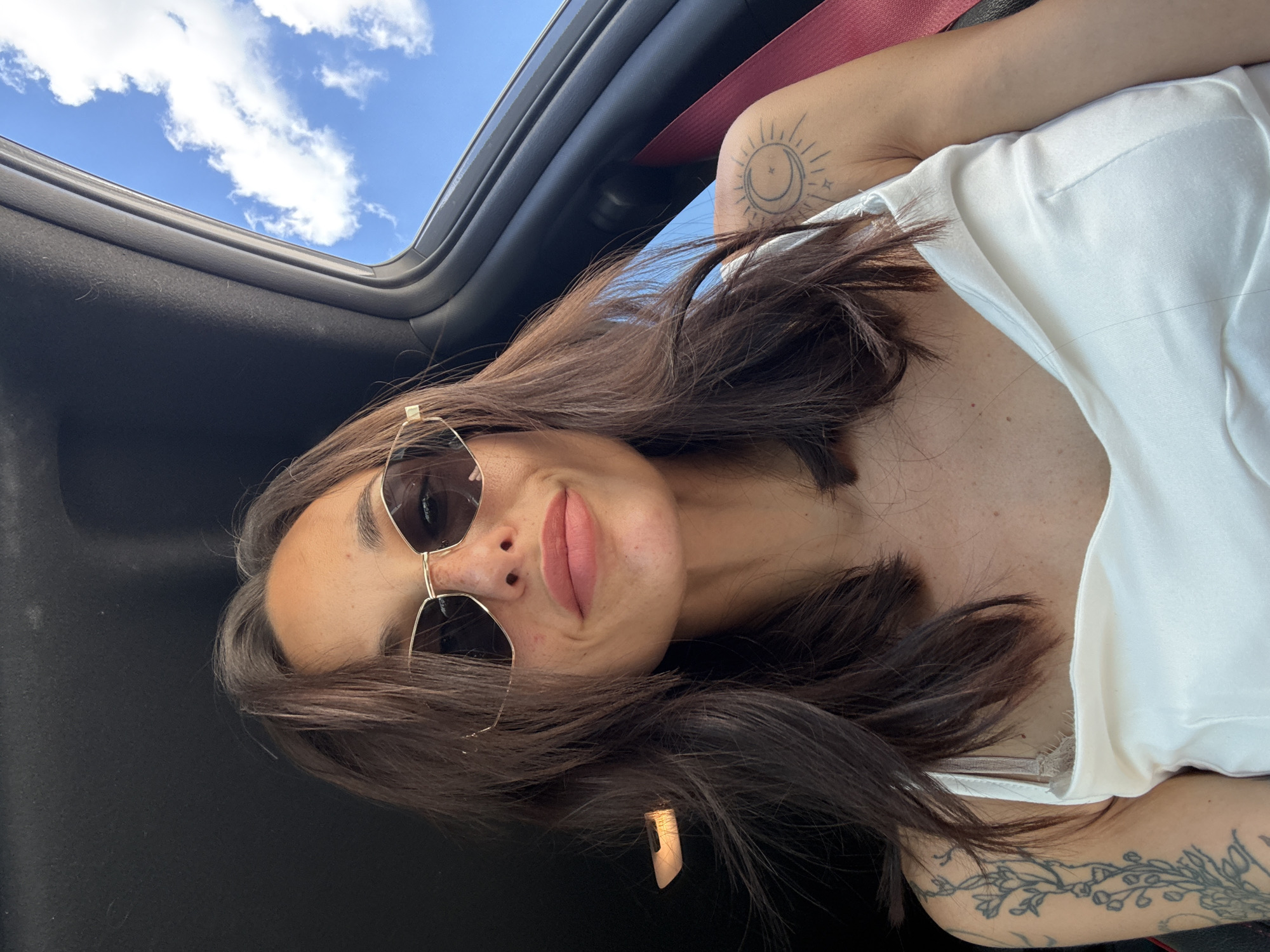 In a selfie taken from inside a car, a young woman with dark brown hair and sunglasses looks into the camera. She is wearing a white tank top and has tattoos on her left arm and shoulder. The car's sunroof is open, revealing a bright blue sky with white clouds. The photo appears to have been taken during the day. There is no indication of the specific location in Pleven, Bulgaria.