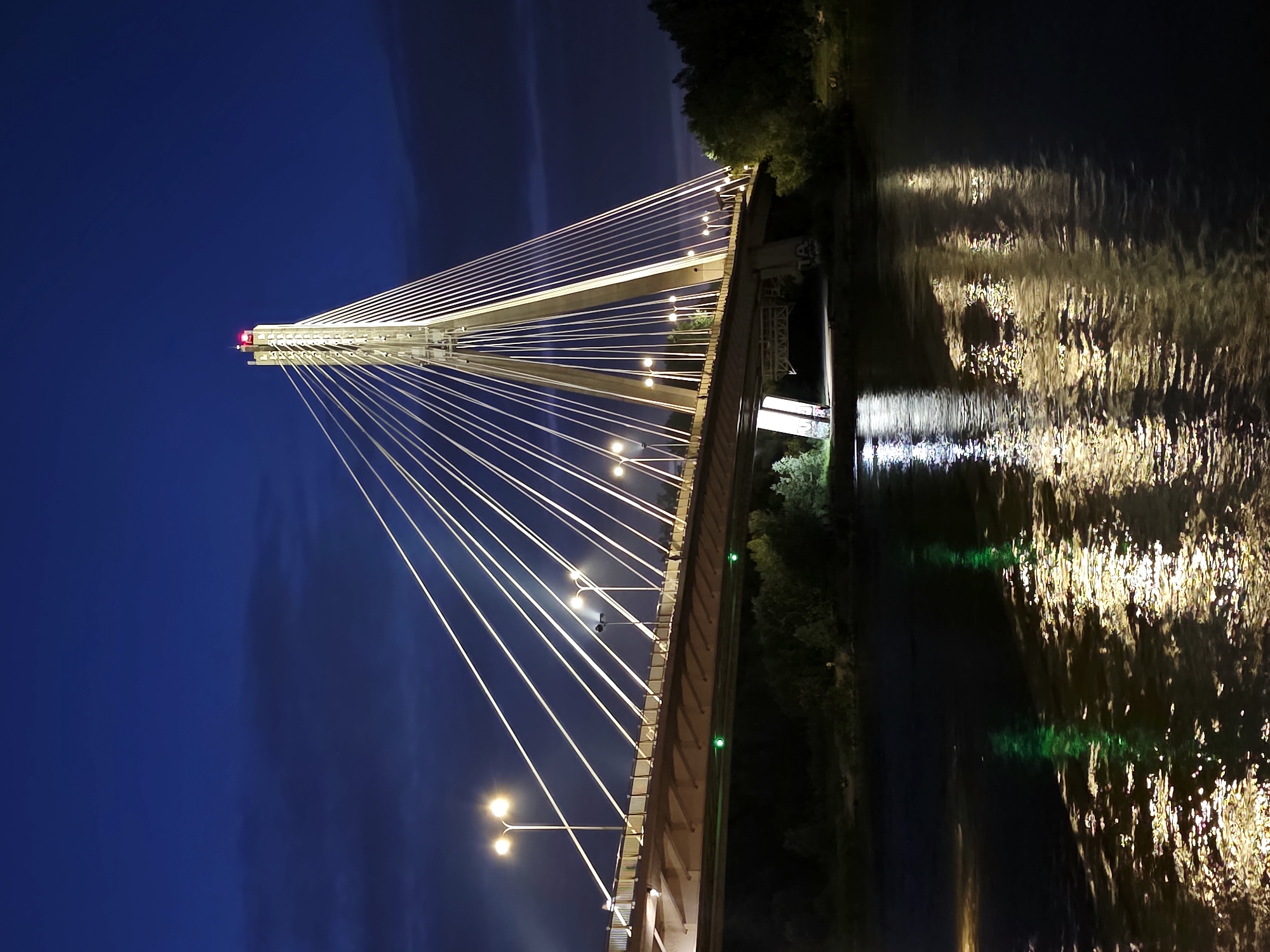 The image depicts the Skrapar Bridge in Warsaw, Poland at night. The bridge is a cable-stayed bridge illuminated by streetlights and lights on its cables and tower. The tower is tall and slender, with a red light at its apex. The bridge spans a body of water, likely the Vistula River, with its lights reflecting on the dark surface. Trees line the riverbank. A few people can be seen walking on the bridge. The sky is a deep blue, indicating it is nighttime.