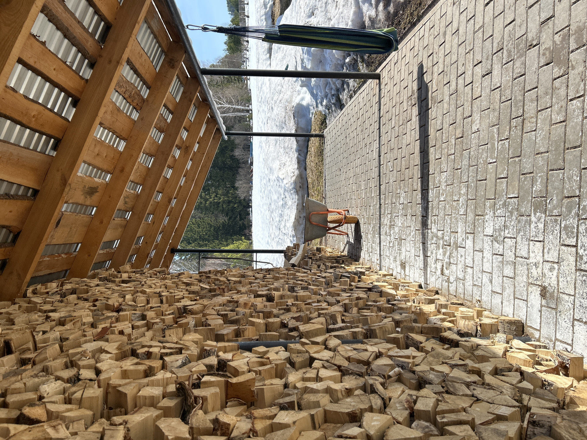 The image shows a large pile of stacked firewood under a wooden shelter structure with a corrugated metal roof. Outside the shelter, a paved area is visible, leading to a partially snow-covered ground and a hammock suspended between posts. A wheelbarrow sits on the edge of the paved area near the firewood. The background includes a treeline and a blue sky. The location is likely Уральское, Russia, based on the user's request. The scene suggests a rural or suburban setting where firewood is used for heating.