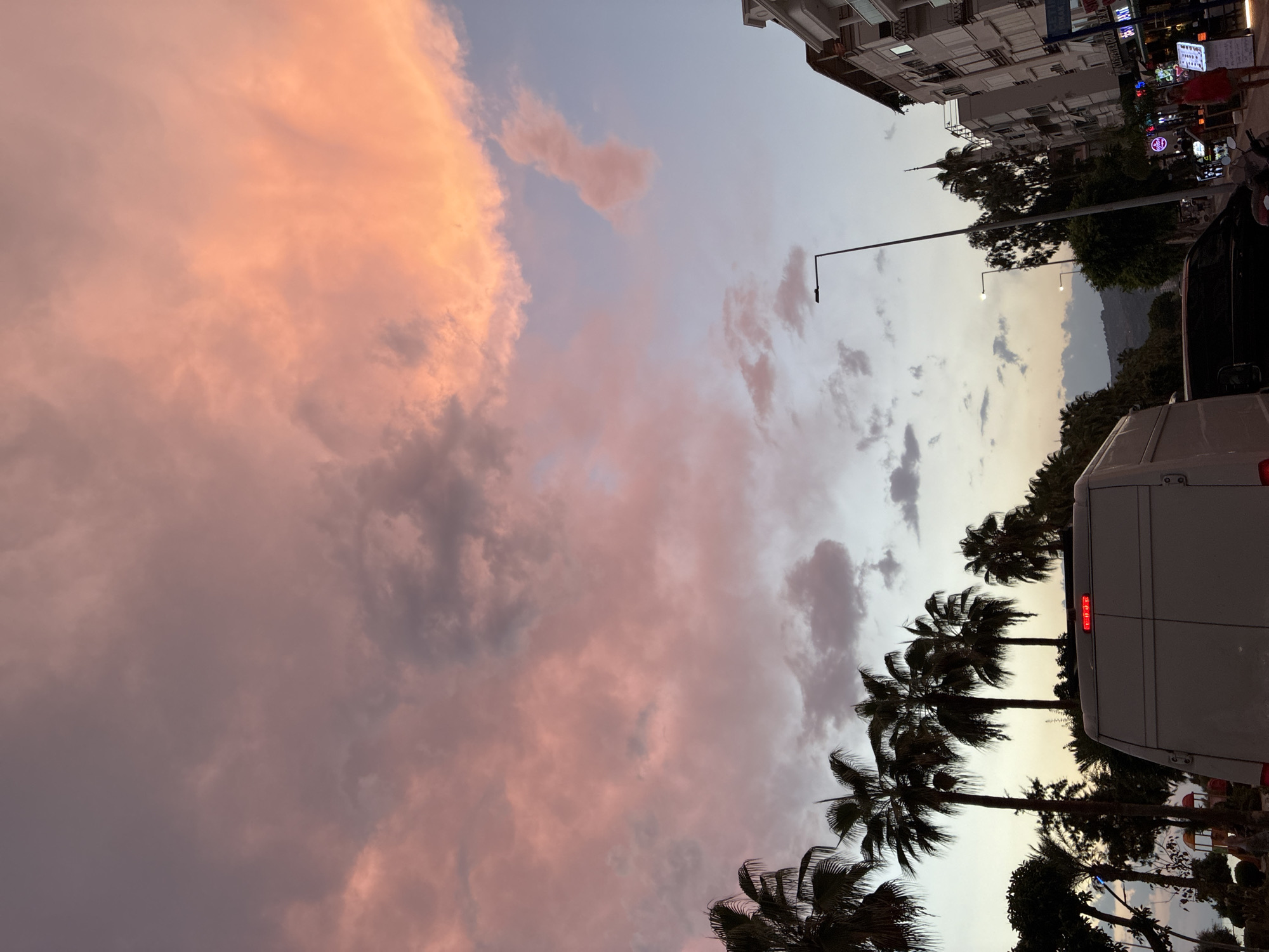 The image captures a view of a street in Alanya, Turkey, during a sunset. The sky is filled with dramatic clouds tinged with shades of pink and orange, indicating the setting sun. Palm trees are visible on the left side of the frame, their fronds silhouetted against the colorful sky. In the foreground, the back of a white van is prominent, with its brake lights illuminated. A black vehicle is parked behind it. The right side of the image shows buildings lining the street, with awnings and signs visible. A street lamp stands tall in the middle ground. Further down the street, more buildings and trees are present, with a distant hillside visible. There are a few individuals walking on the sidewalk on the right, and various shop signs are illuminated.
