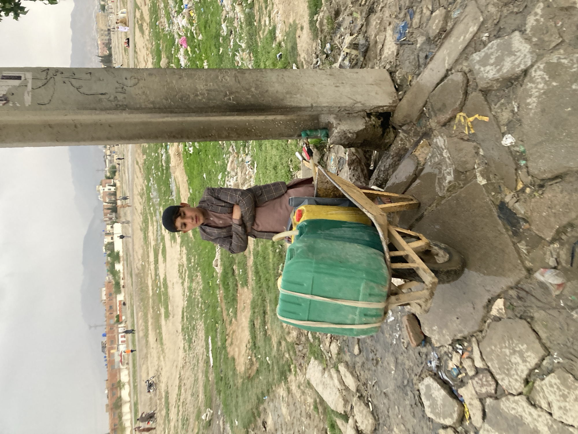 A young boy with dark hair and a brown plaid jacket stands next to a wooden cart carrying stacked green and yellow containers. The cart is positioned on a broken concrete sidewalk alongside a pole. The surrounding environment appears to be a dirt and grass area littered with trash. In the distance, a mountainous landscape and city buildings are visible. The location is identified as Quetta City Tehsil, Pakistan.