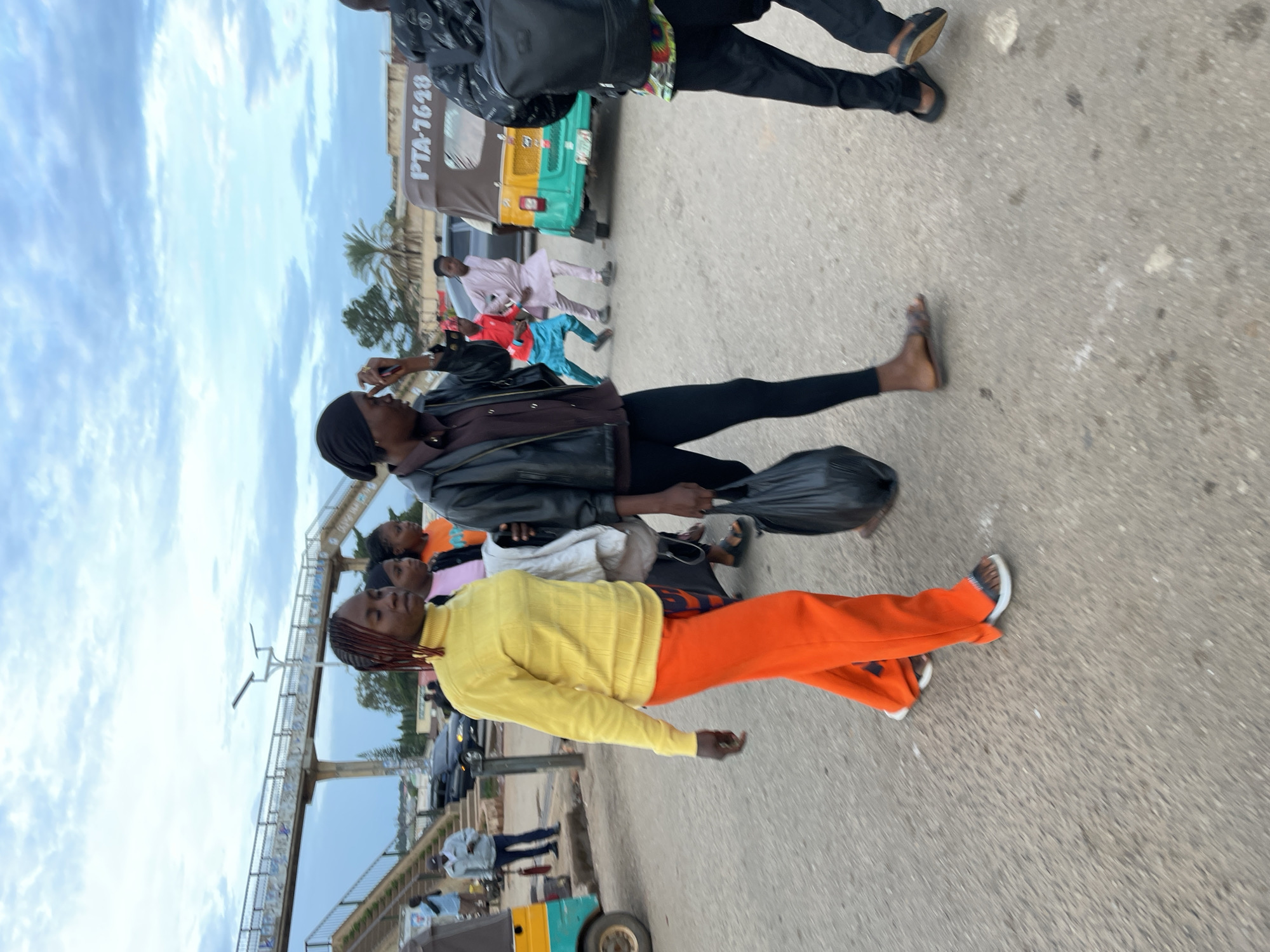 The image depicts an outdoor urban street scene in Gura Top, Nigeria, under an overcast sky with scattered light clouds. The ground is an asphalt or concrete road surface.
In the foreground, from left to right, three individuals are walking towards the right side of the frame. The leftmost individual is a woman with braided hair, wearing a yellow turtleneck sweater, bright orange wide-leg pants, and dark-strapped sandals with white soles. Her left hand holds a light-colored garment. The central individual is a woman wearing a black head covering, a dark brown collared shirt, a black leather jacket, black leggings, and dark sandals. She holds a black plastic bag in her left hand, and her right hand is positioned near her temple. Partially visible to her right is a third individual, seen from the back, wearing a black backpack and black pants.
In the midground, behind the foreground individuals, two women are partially visible: one wearing a pink top and another wearing an orange top. Further back, on the left, a man in a light-colored shirt and dark pants stands near a yellow and teal three-wheeled vehicle, partially visible. A second man in a light blue shirt and dark pants stands further left near a sidewalk. In the midground right, a yellow and green three-wheeled auto-rickshaw (keke Napep) with the identifier "PTA-7648" on its rear is visible, with a partially obscured Nigerian license plate below it. To the left of this vehicle, two men walk; one wears a red top and blue pants, and another wears a light pink traditional garment.
In the background, a concrete pedestrian bridge with a patterned railing and multiple sets of stairs spans across the upper-left portion of the scene, featuring two streetlights on poles. Distant buildings, various trees, and palm trees are visible under the horizon line. A dark-colored sedan is also visible on the street in the background.