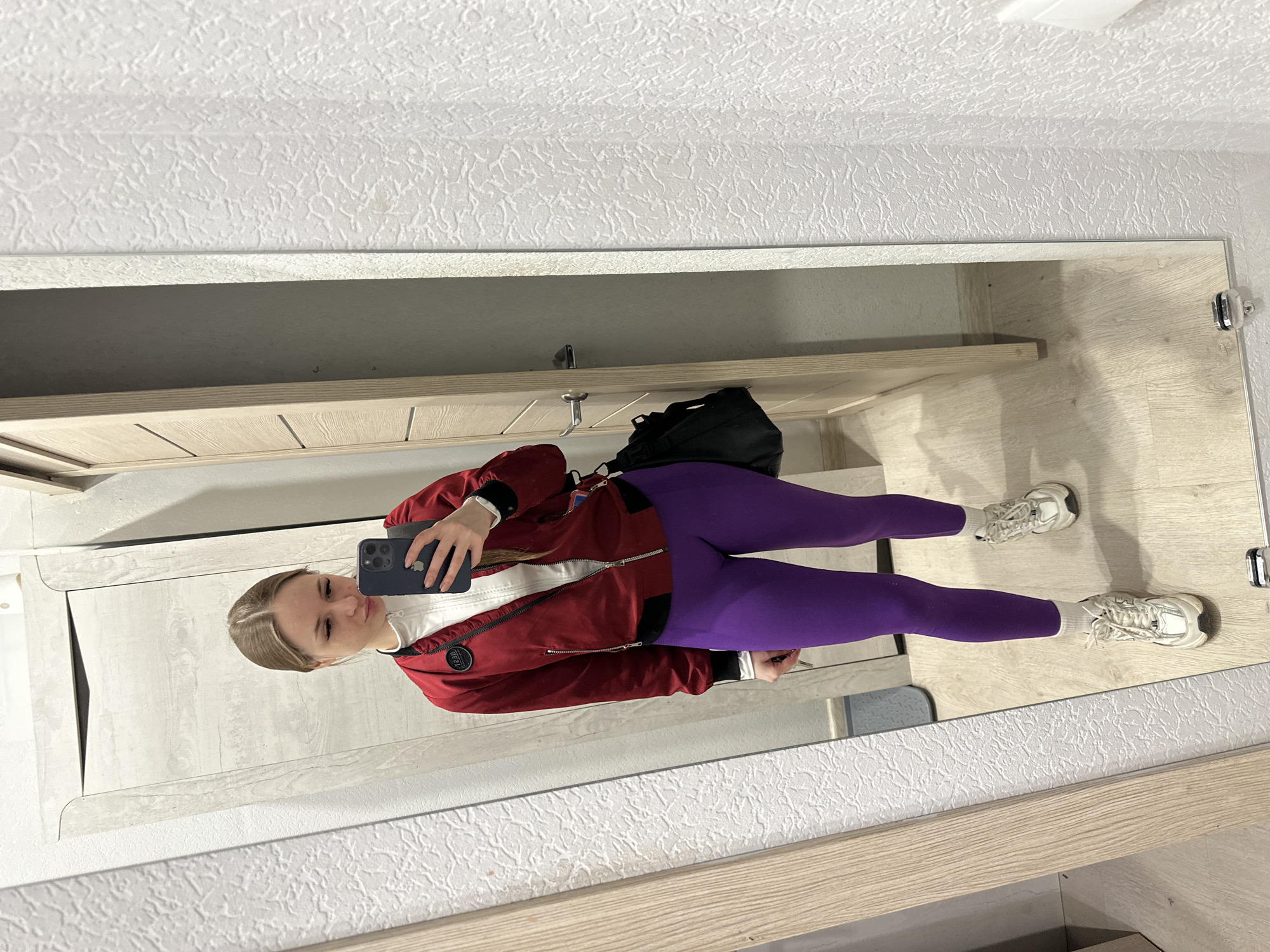 A mirrored image of a young woman taking a selfie with her phone. She is wearing a white top, a red jacket, purple leggings, white socks, and white sneakers. A black bag hangs on the rack above her. The location appears to be indoors, potentially in a hallway or room with a mirrored wardrobe or closet. The walls are white with a textured pattern, and the wardrobe is made of light-colored wood. The location in the city of Unknown, Unknown is indeterminable.