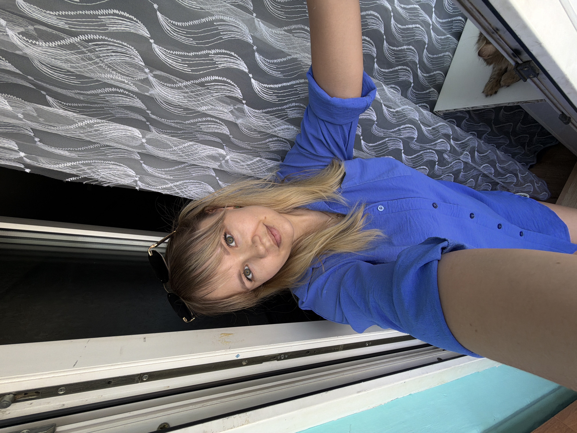A woman in a blue collared shirt takes a selfie inside what appears to be a window frame. Part of her body extends outside the window. The background includes a gauzy, patterned curtain and a small dog resting on a surface outside the window. Sunglasses are placed on top of her head. The photo was taken in Chaikovsky, Russia.