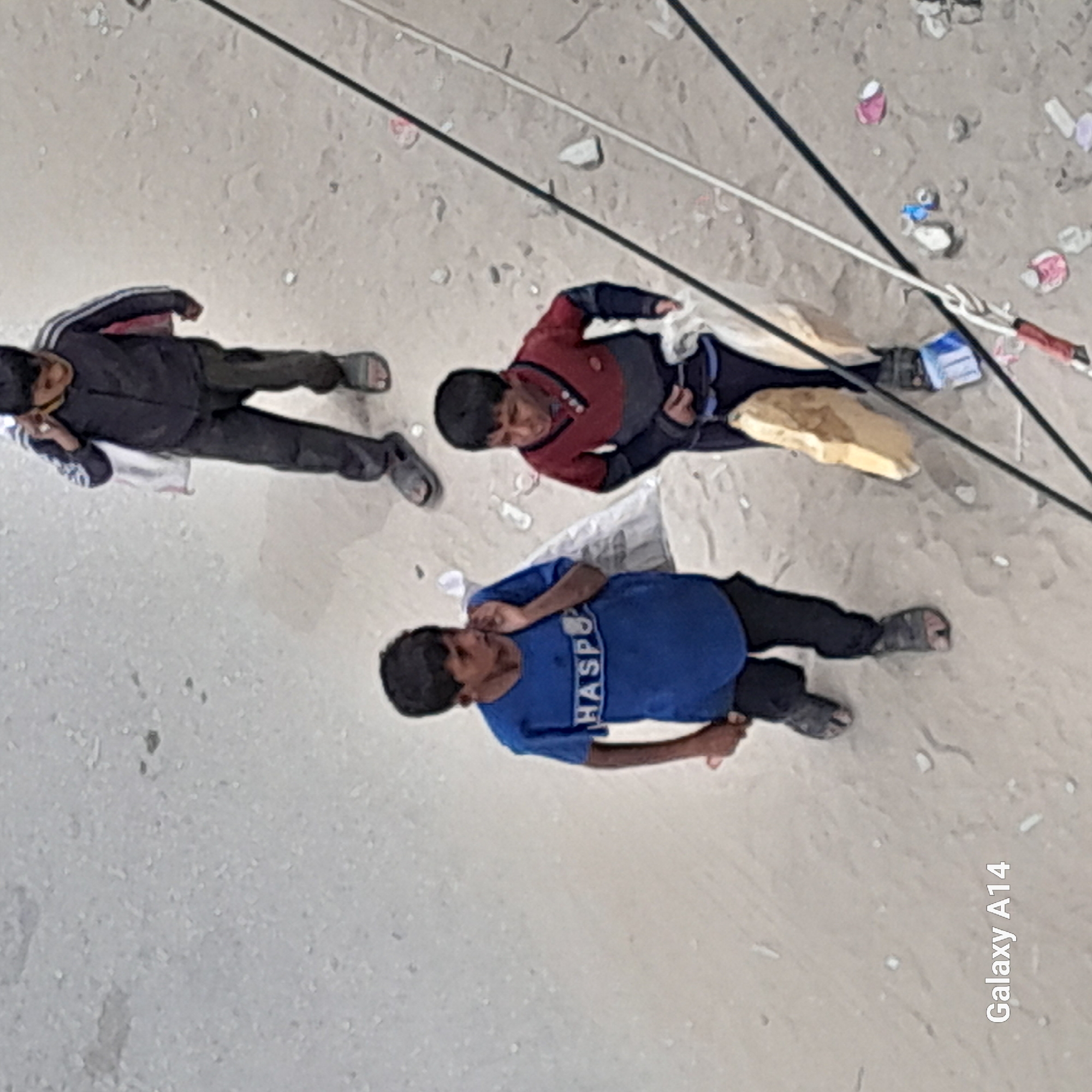 Here's a detailed description of the image:
**Scene:**
The image captures three children lying on a sandy or concrete ground littered with debris. Wires or cables diagonally cross the scene in the upper right.
**People:**
* **Child 1:** Lies on the left in a dark jacket with white stripes on the shoulder, dark pants, and sandals.
* **Child 2:** Lies in the middle with a red shirt with grey sleeves, dark pants, and sandals.
* **Child 3:** Lies on the bottom with a blue t-shirt that says "HASPO" on it, dark pants, and sandals.
**Objects:**
* Debris scattered on the ground (e.g., cans, wrappers).
* Wires or cables overhead.
* Plastic bags.
* Piece of cardboard.
* Sandals.
**Actions/Interactions:**
* The children are lying down on the ground.
**Environment:**
* The scene is an outdoor area with a sandy or concrete ground. There is litter present, suggesting an urban or industrial environment.
**Location:**
* Unknown, Unknown