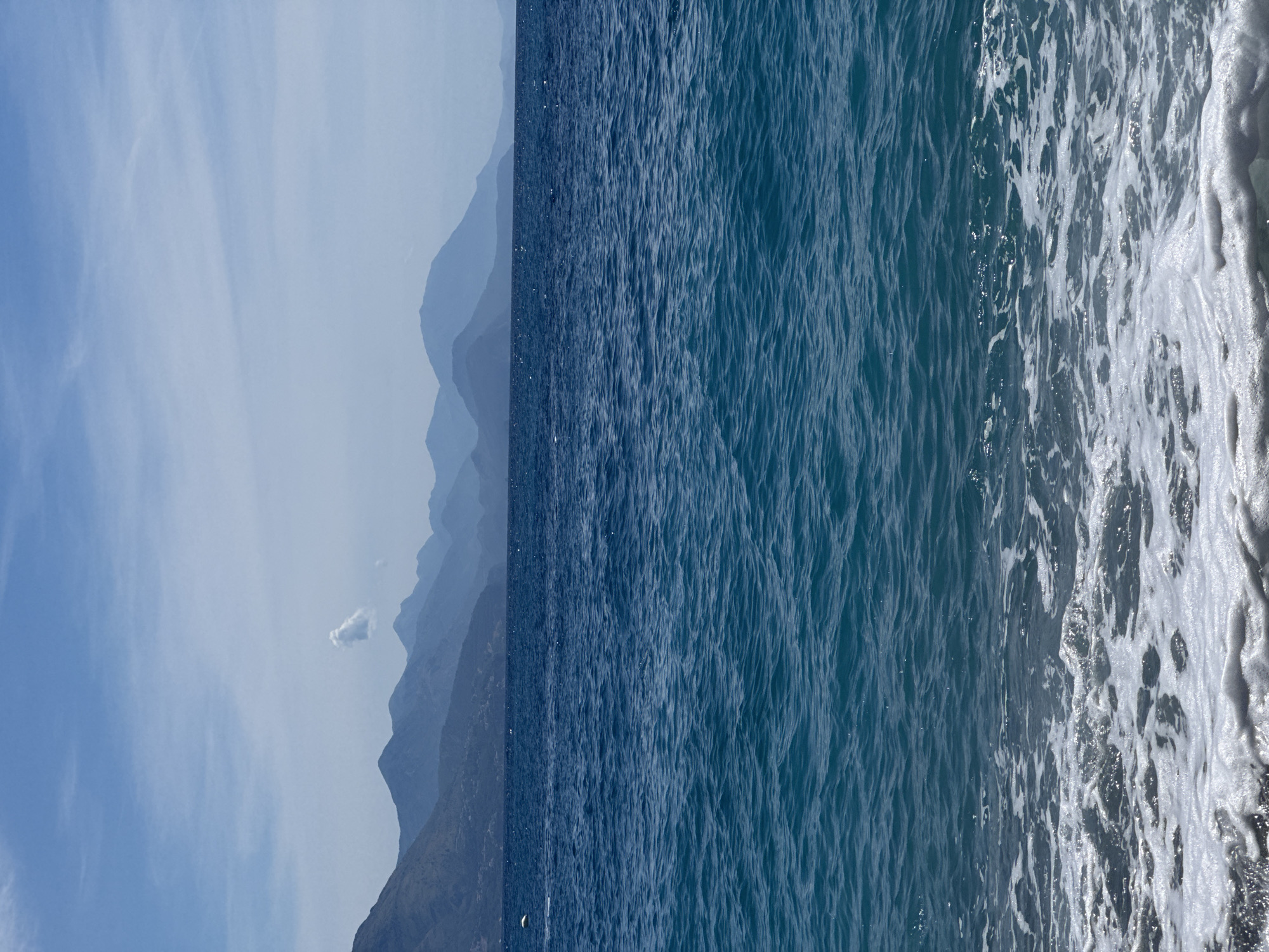 A serene coastal scene unfolds, dominated by the expansive blue of the Adriatic Sea meeting a distant, misty mountain range. The water in the foreground is a vibrant turquoise, rippling gently and capped with frothy white waves as they wash ashore onto a pebble beach. The sea stretches out to the horizon, where a line of layered mountains, rendered in soft hues of blue and purple due to atmospheric perspective, dominates the background. The sky above is a pale, clear blue, with a few wispy clouds scattered across its expanse, one notably shaped like a puff of smoke. The overall impression is one of tranquility and natural beauty, capturing a peaceful moment in Prijevor, Montenegro. There are no people or specific man-made objects visible in this natural landscape.