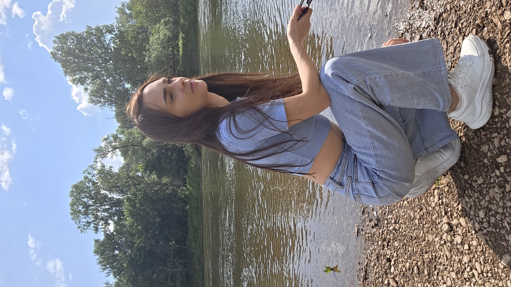 A young woman with long brown hair is crouching by the water's edge on a rocky bank, looking to her right. She is wearing a light blue, short-sleeved crop top and light blue wide-leg jeans, along with white sneakers. The water is a calm river, with trees and foliage lining the opposite bank under a bright blue sky with scattered clouds. The location appears to be a natural, undeveloped area, consistent with the rural setting of Дарьино, Russia.