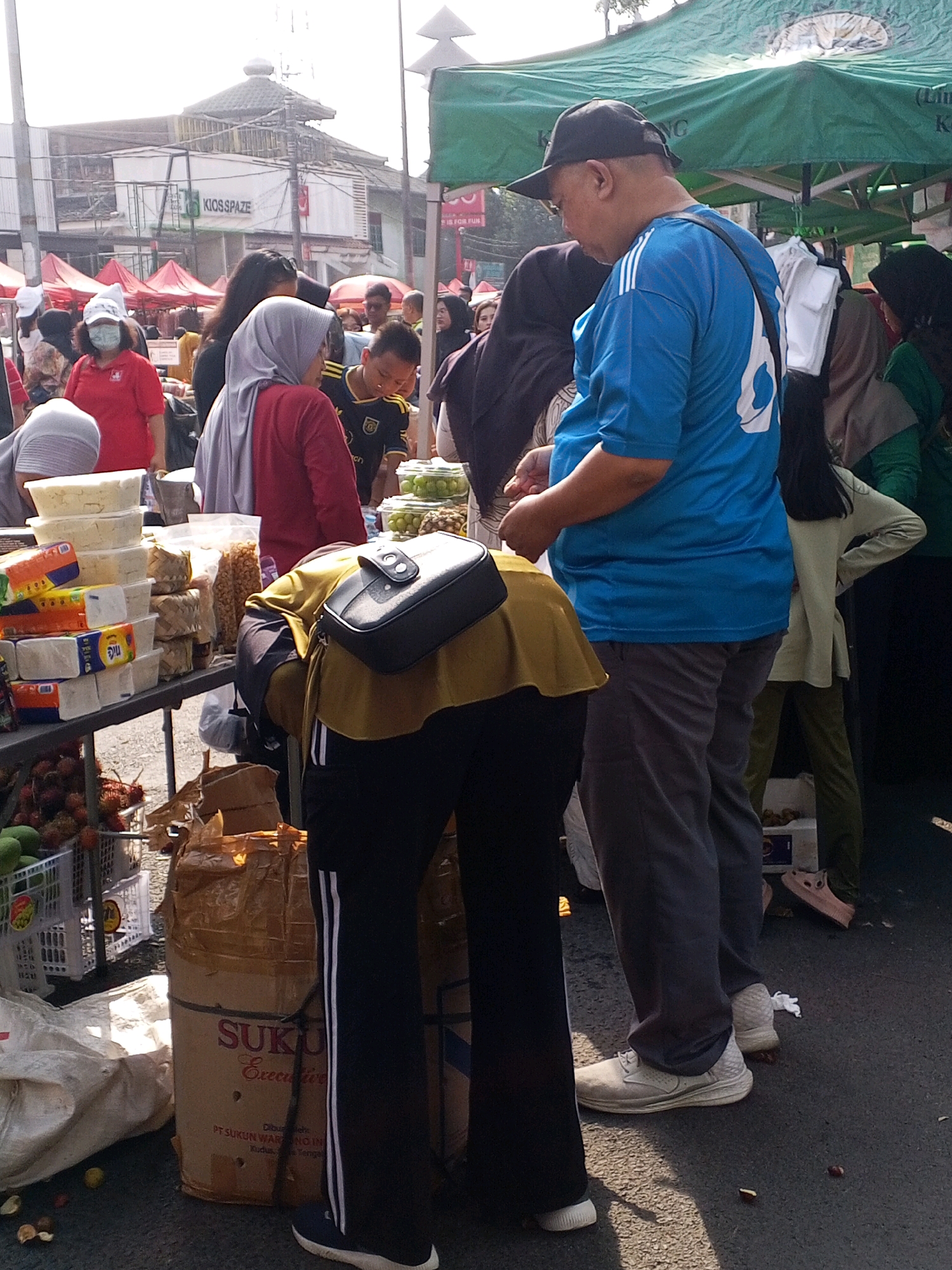 The image captures a bustling street market scene, likely in Serang, Indonesia, judging by the context of its origin. The foreground features a stall laden with various food items, including what appear to be blocks of cheese or tofu and bags of snacks. In the center, a person in a yellow top and black pants with white stripes is bending over, obscuring their face. To their right, a man in a blue t-shirt with a large white number '6' on the back and a black baseball cap is looking down. Behind them, a green tent provides shade over a display, possibly of produce. Numerous other individuals, many wearing headscarves, are visible in the background, suggesting a busy market environment. Red market tents are visible further back, along with buildings and power lines, indicative of an urban setting. The ground is paved, and there are various bags and containers scattered around, typical of a market area.