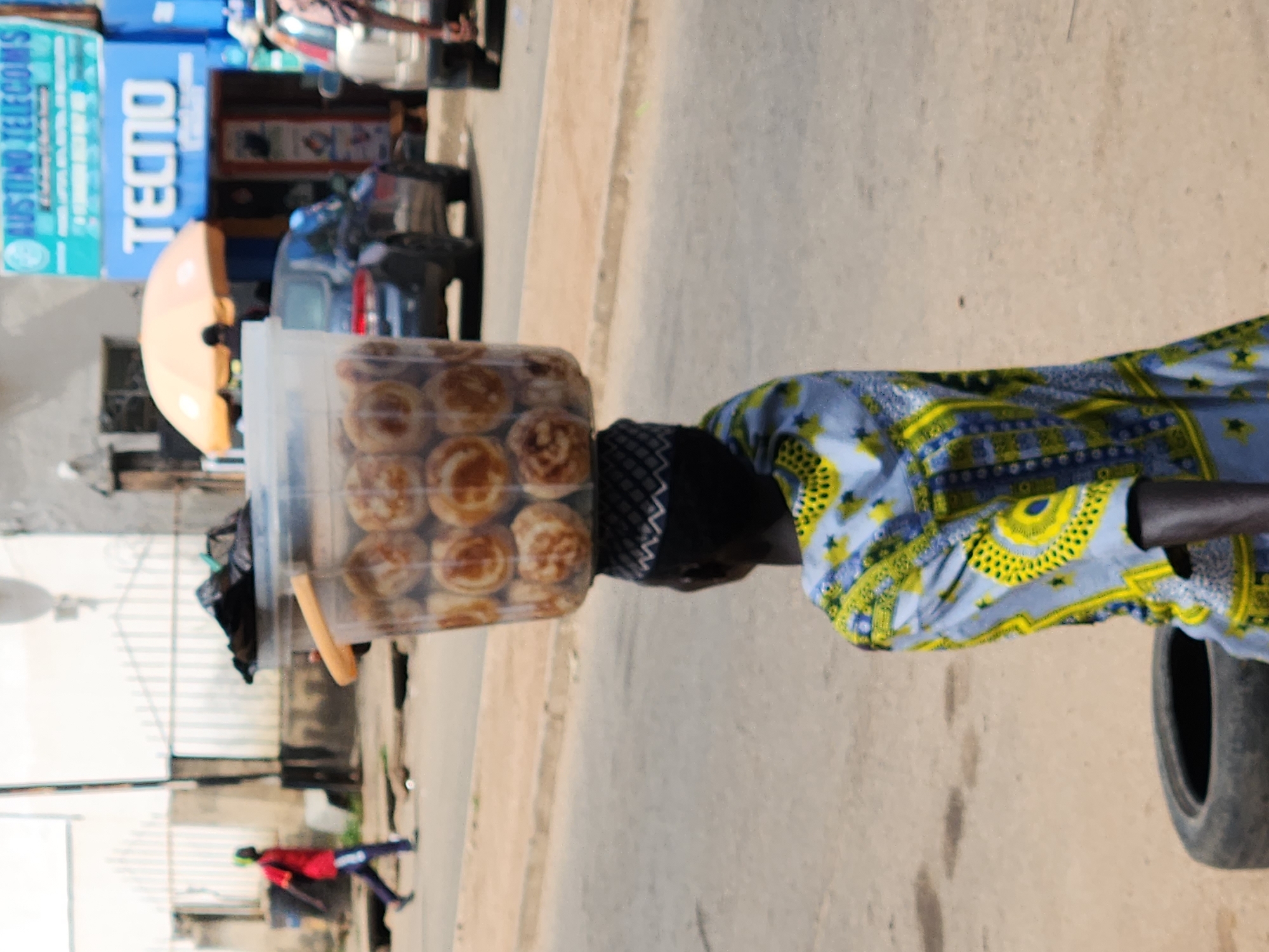 A person is walking away from the camera in the city of Jos, Nigeria, with a large bucket of food balanced on their head. The person is wearing a vibrant blue and yellow patterned garment and a dark patterned cap. In the foreground, a tire lies on the dusty road. In the background, a black car is parked on the side of the road, and further in the distance, buildings with signs indicating "TECNO" and "AUSTINO TELECOMS" are visible. Another person in a red shirt and dark pants walks in the opposite direction on the sidewalk. An orange umbrella is also visible in the background.