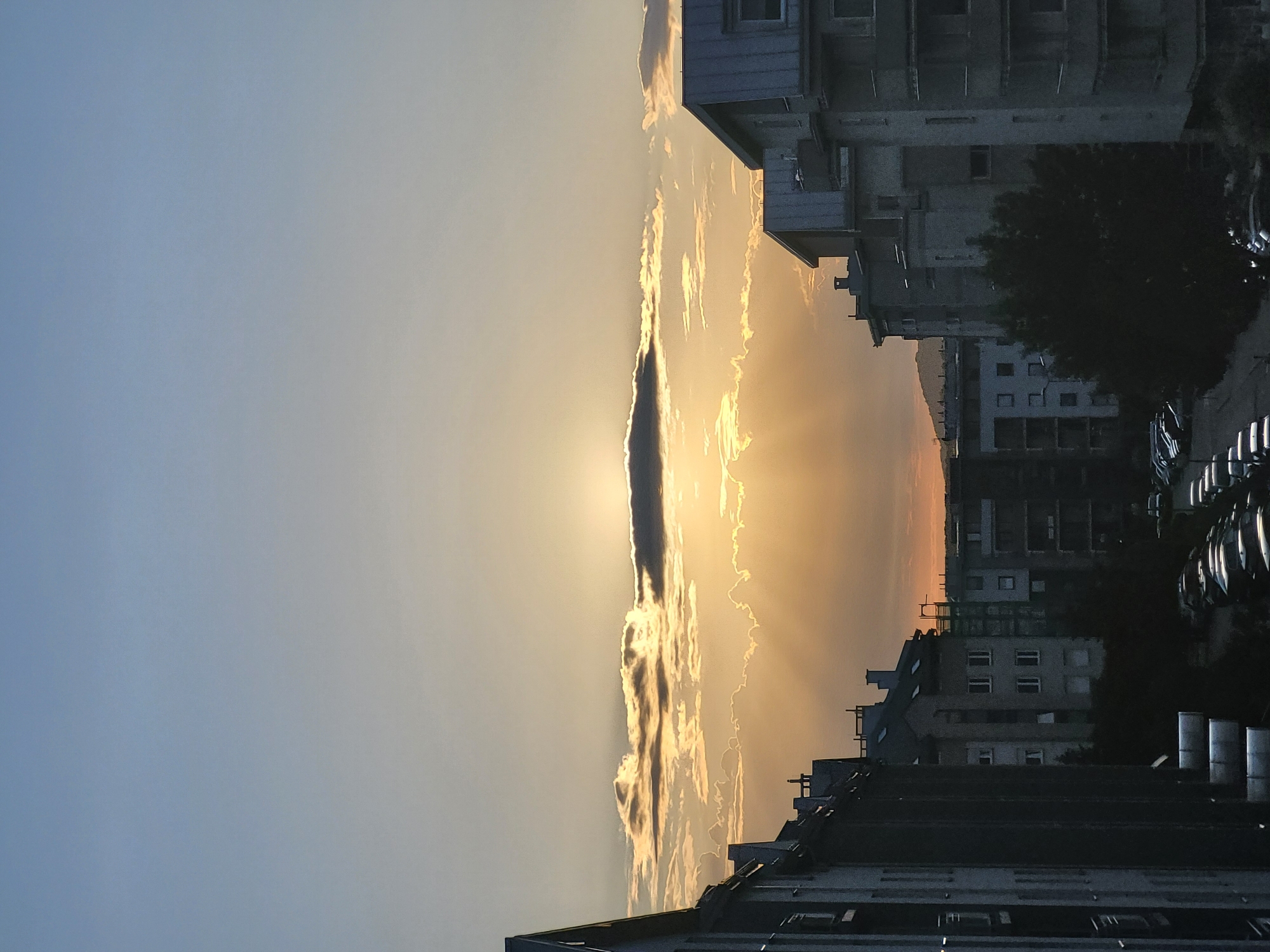 The image depicts an urban landscape at either sunrise or sunset, in the city of Unknown, Unknown. A low sun is largely obscured by a prominent, dark, elongated cloud positioned horizontally across the mid-sky. Bright crepuscular rays emanate downwards from behind this cloud, illuminating the lower atmosphere in warm shades of orange and yellow, contrasting with the lighter blue-gray sky above. Scattered, thinner clouds are also visible, some brightly lit, others in shadow.
The foreground and midground are dominated by multiple multi-story buildings, likely residential, with sloped roofs and visible windows. These structures appear largely silhouetted or in deep shadow due to the backlight from the low sun. Buildings on the far left and right frame the central view, with their architectural details, such as balconies, chimneys, and attic windows, partially discernible despite the low light. In the lower center and right, a dense concentration of parked passenger cars fills a street or parking lot. A dark, leafy tree is situated in the lower center, partially obscuring the view of the vehicles. Distant hills or mountains are faintly visible on the horizon line. No people are present or discernible in the scene.